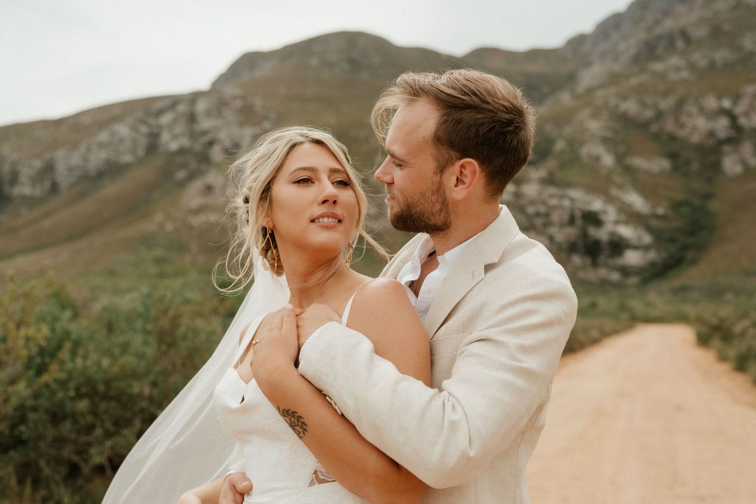 A romantic wedding couple embraces on a dirt road with rugged mountain scenery behind them.