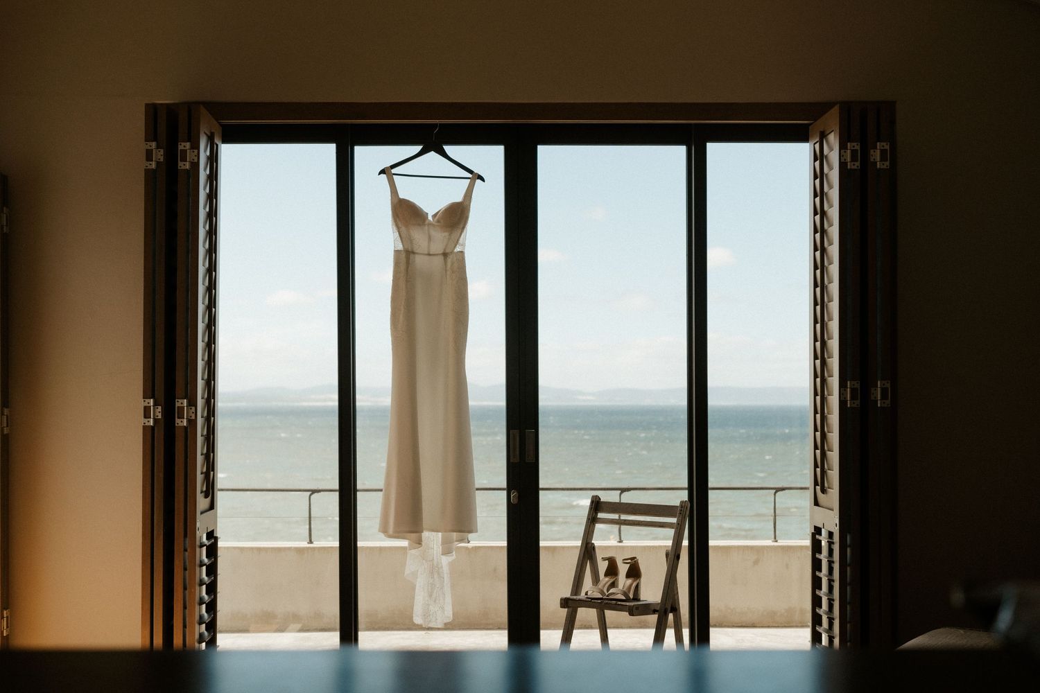 A wedding dress hangs in doorway frame overlooking an ocean view balcony.