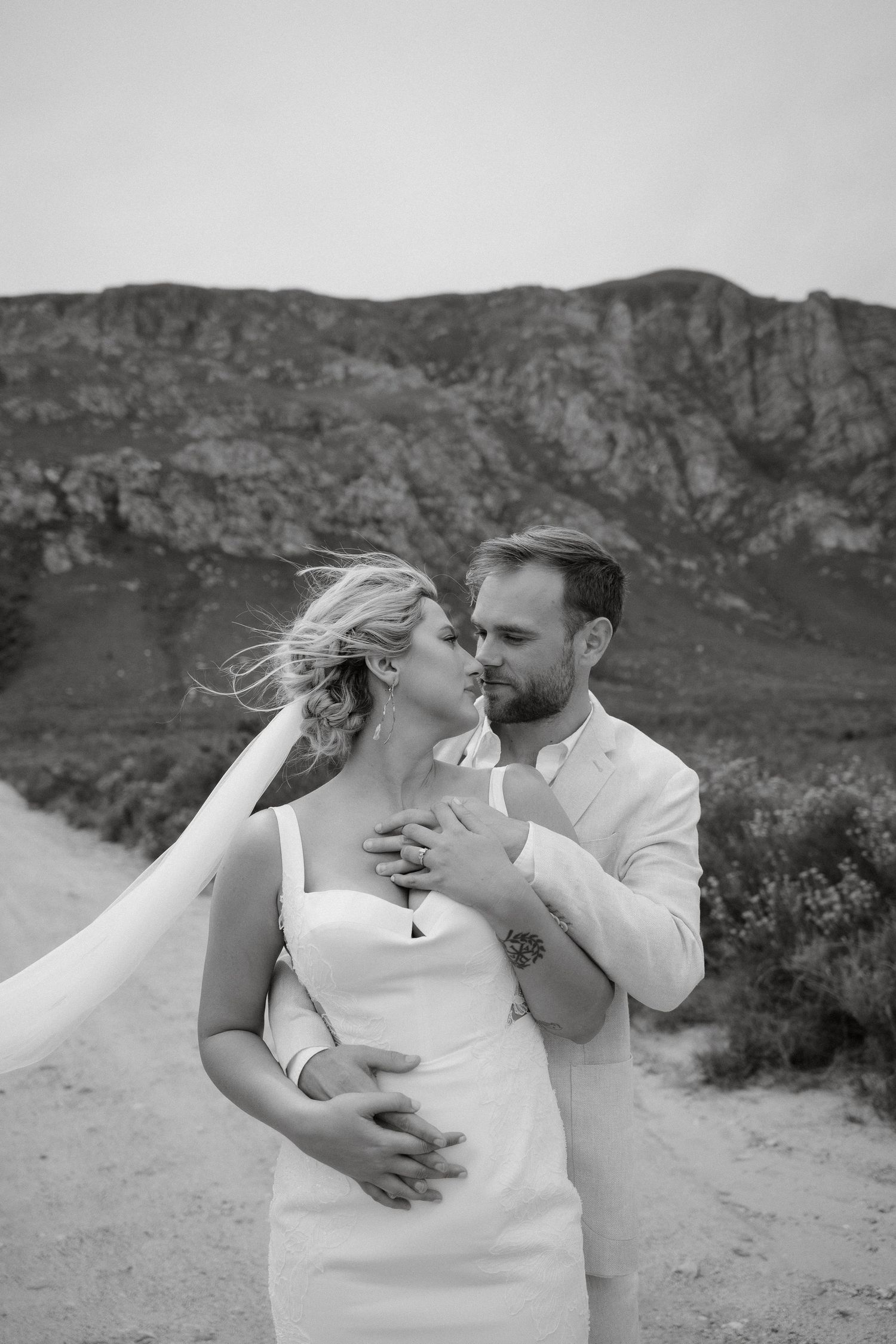 Black and white portrait of newlyweds sharing an intimate embrace against mountainous terrain.