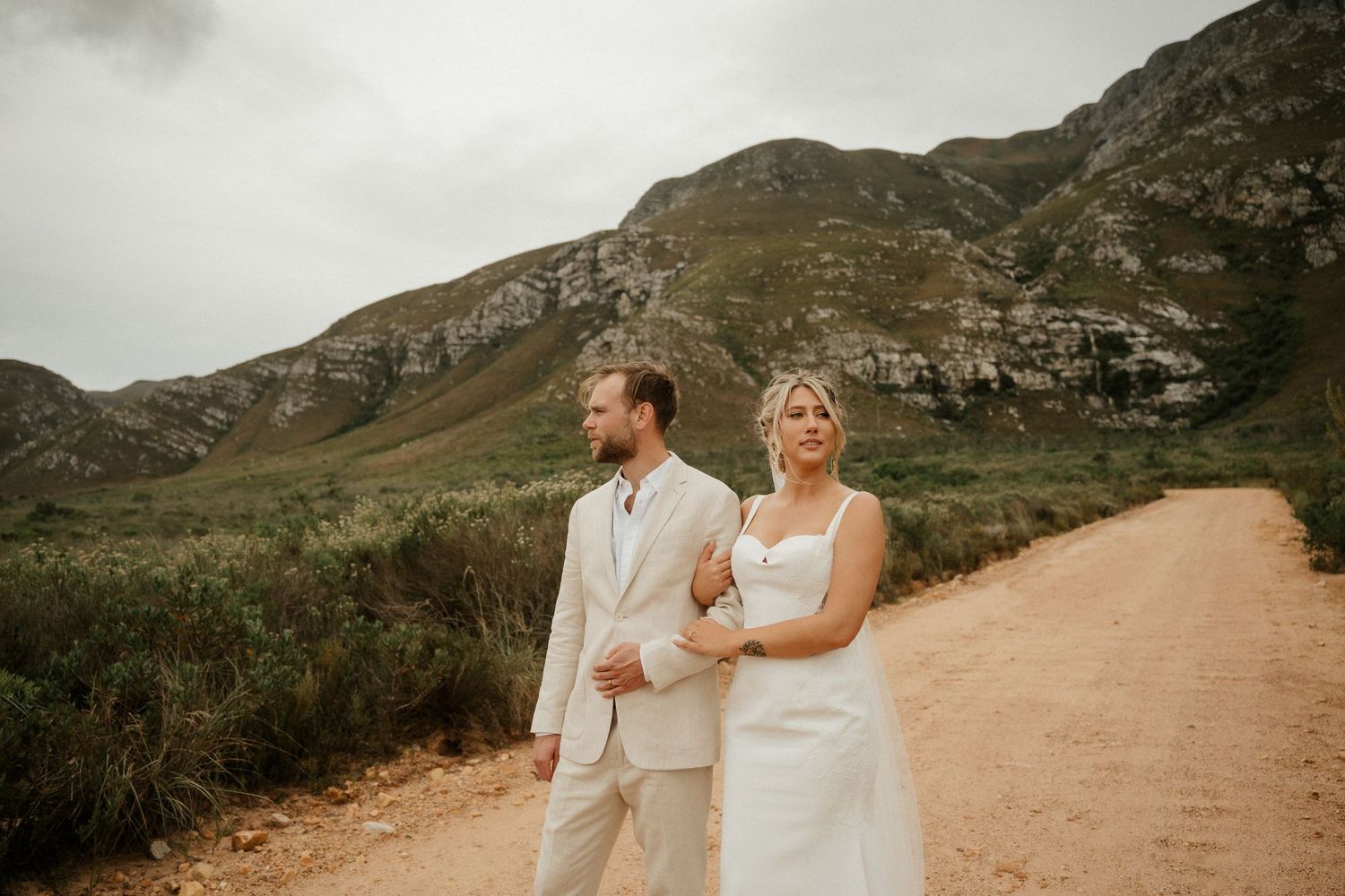Newlyweds pose together on a scenic mountain trail with dramatic cliffs in the background.