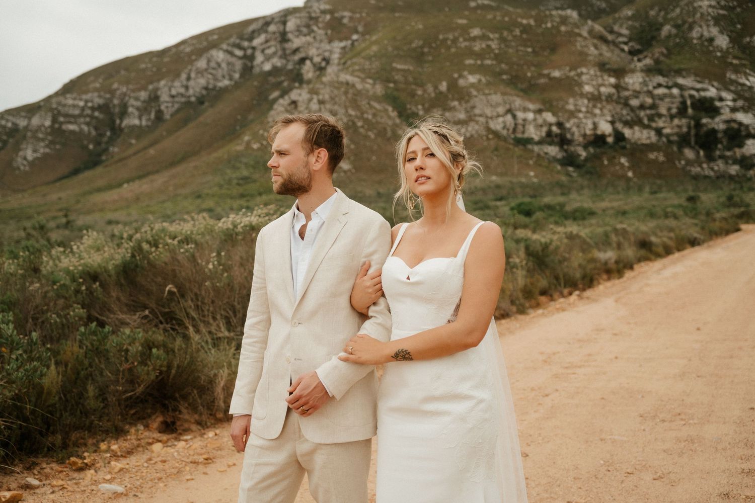 A wedding couple in light-colored attire walking along a mountain path together.