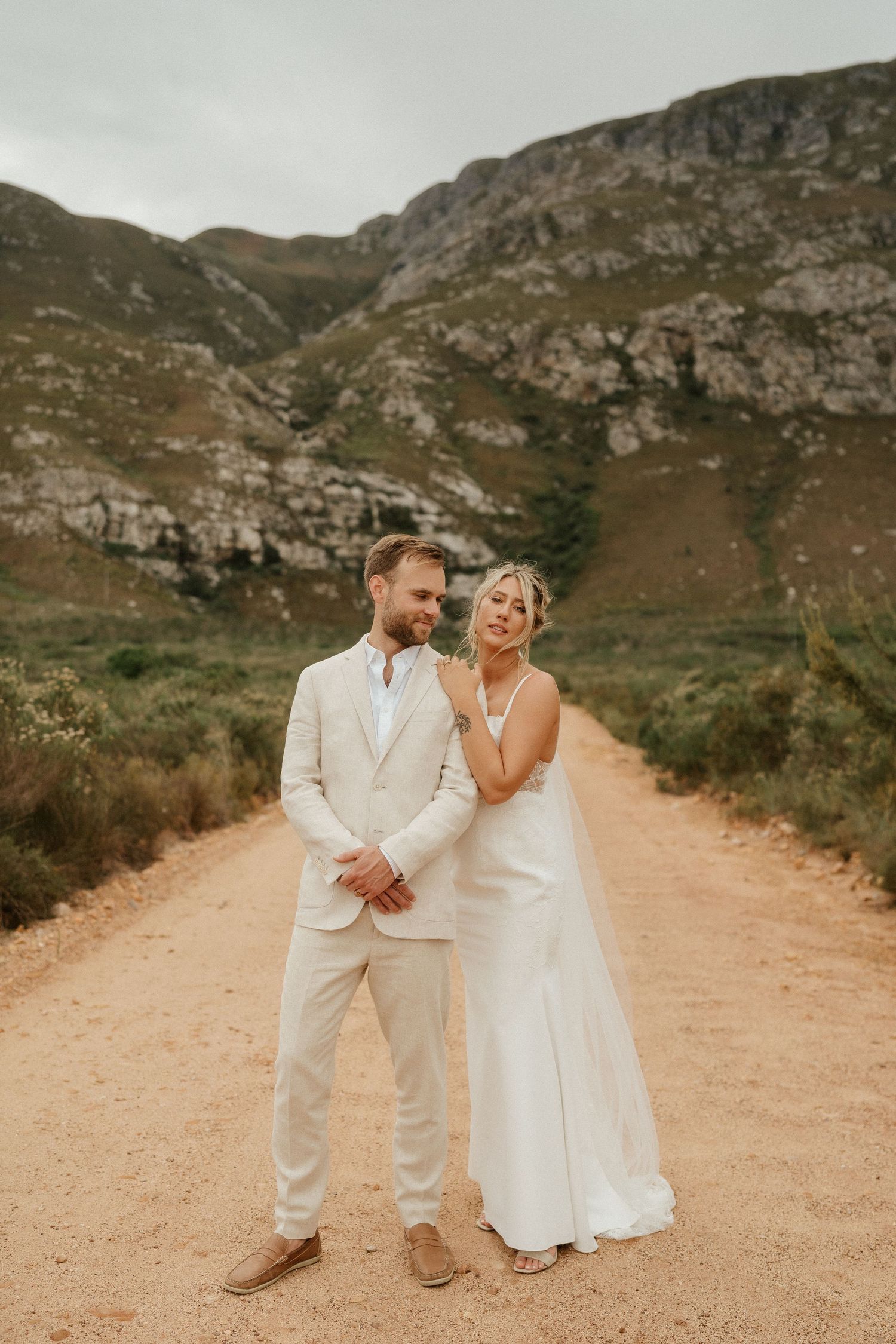 Wedding couple in elegant white attire share a playful moment on a dusty mountain path.
