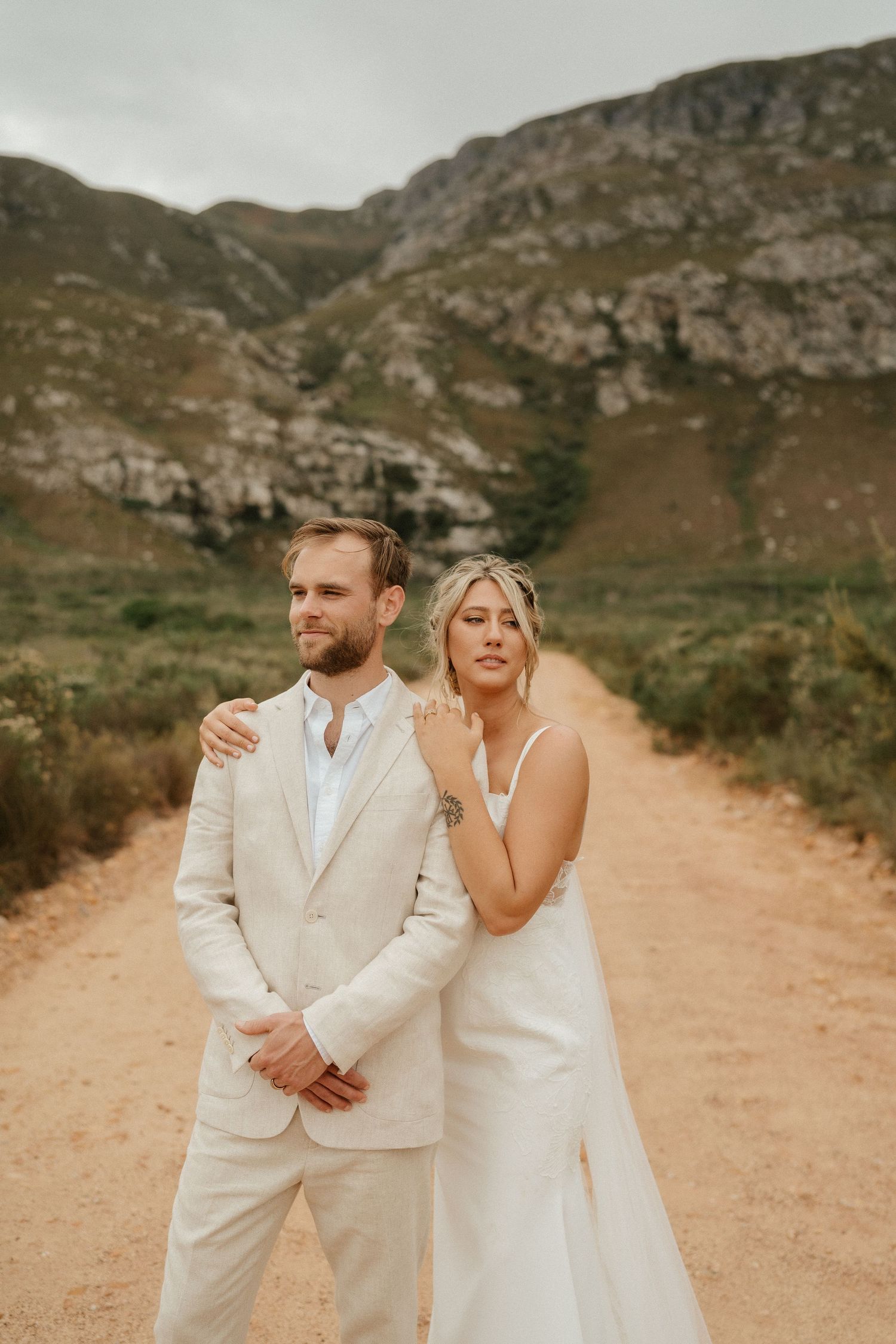 A wedding couple poses playfully on a mountain path surrounded by dramatic rocky terrain.