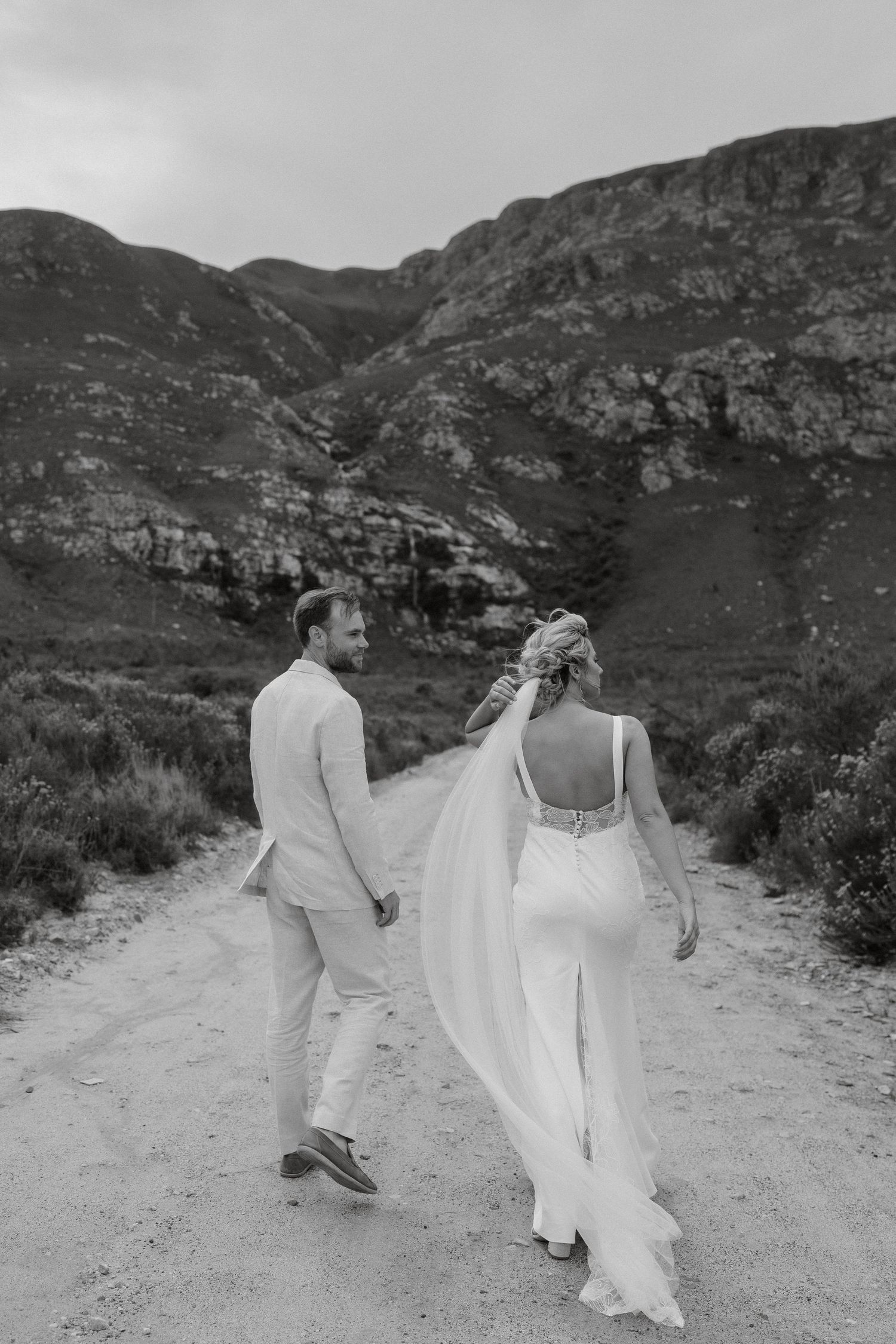A black and white photo of a couple walking hand in hand down a mountain path in wedding attire.