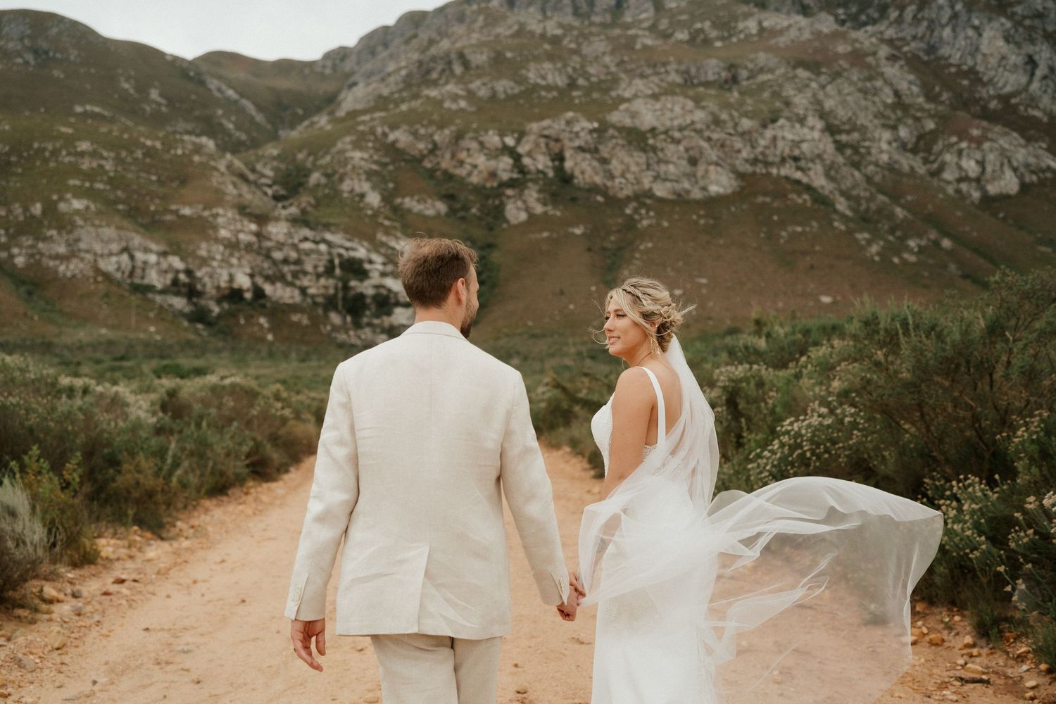 Couple in white wedding attire walking hand in hand on mountain path with flowing veil.