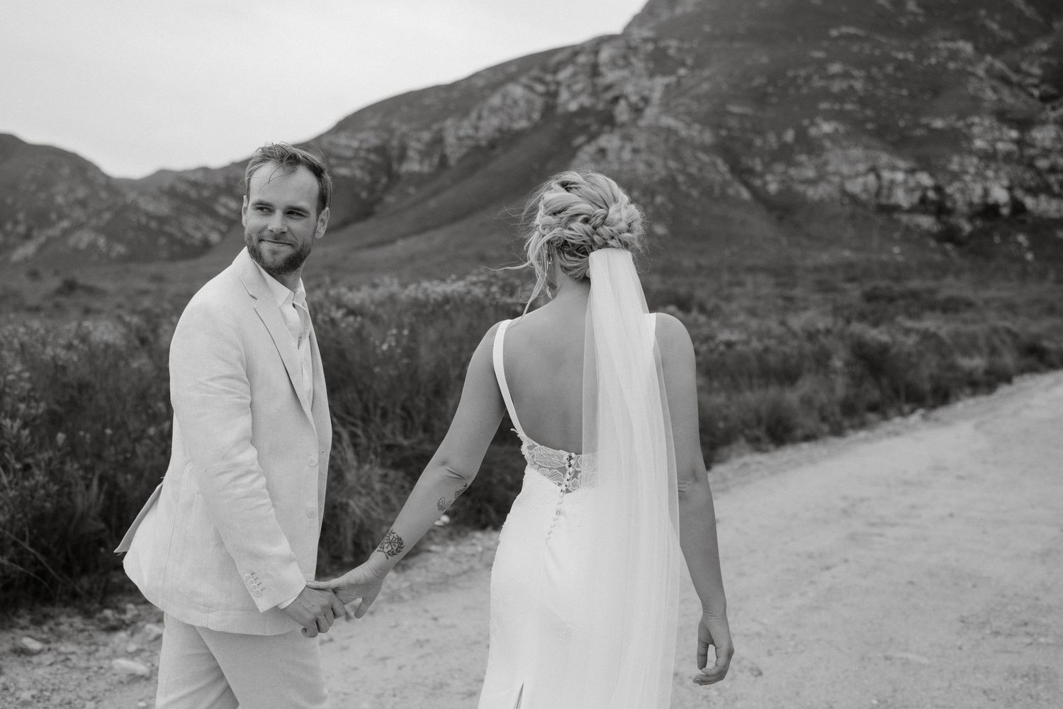 A black and white photo of newlyweds holding hands while walking along a mountain path.