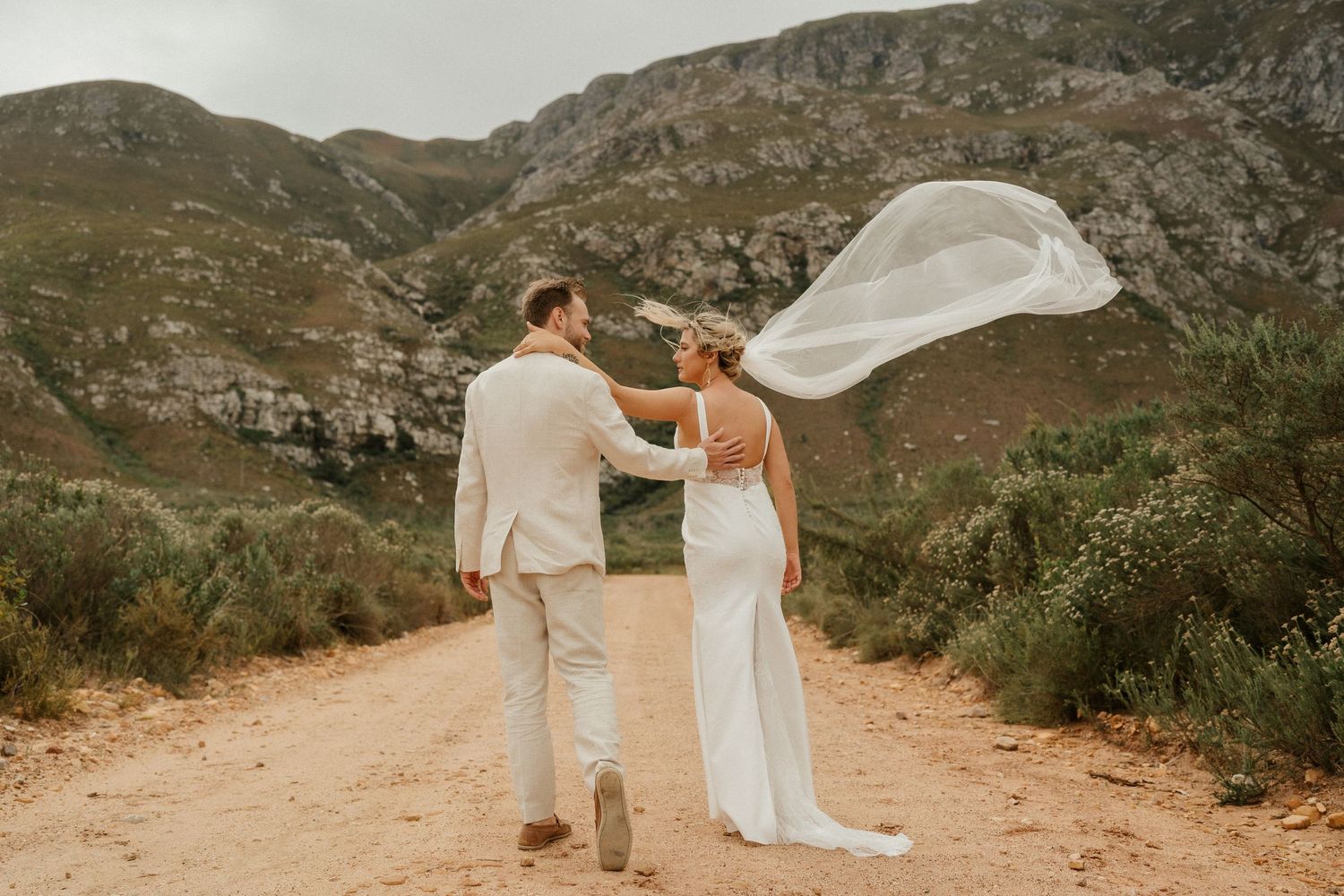 A wedding couple stands on a scenic mountain trail as the bride's veil flows dramatically in the wind.
