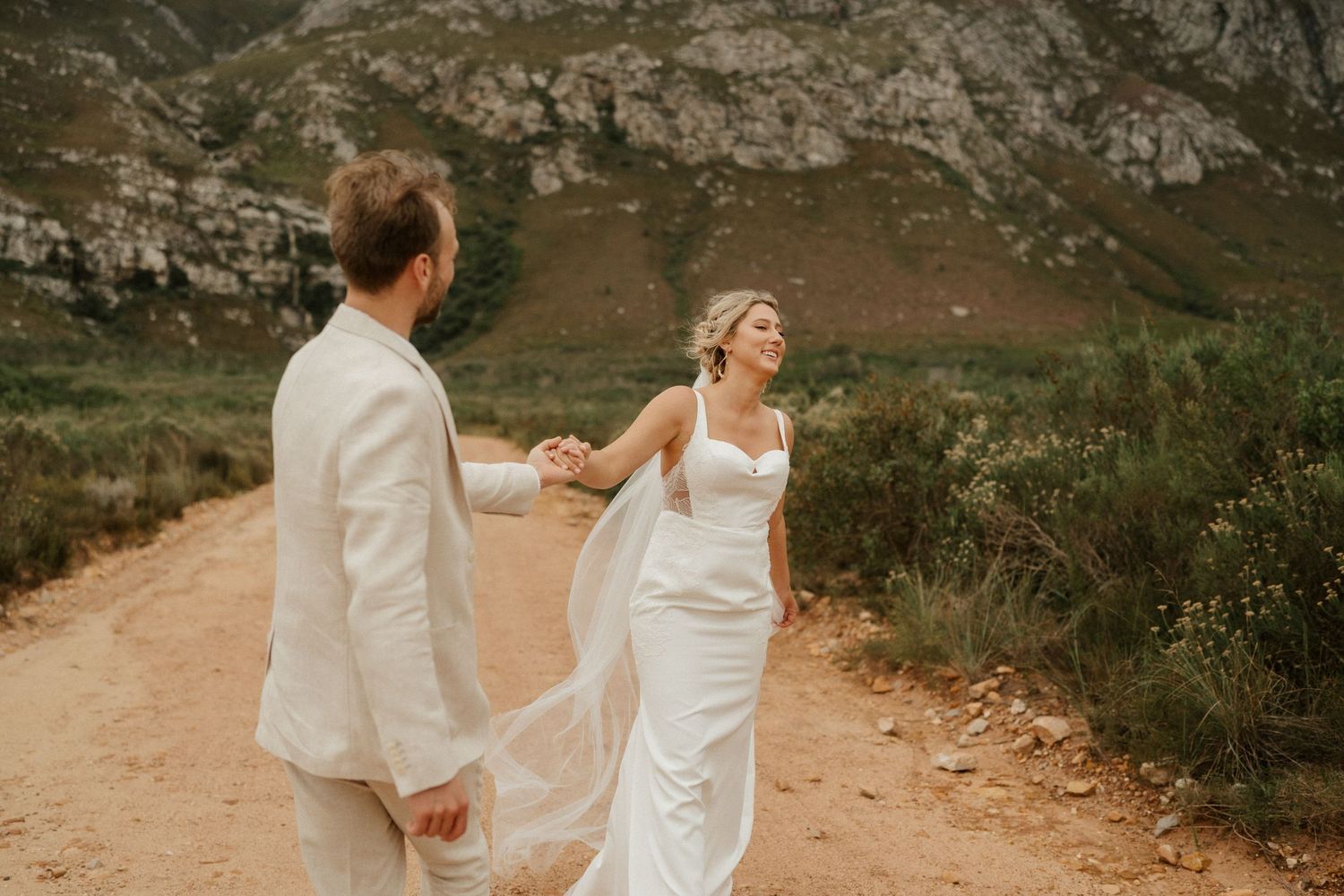 Newlyweds in white attire share a joyful moment on a mountain trail during their wedding photoshoot.