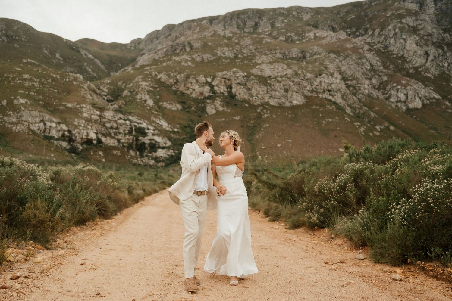 Couple in wedding attire sharing a romantic moment on a dirt path surrounded by mountains.