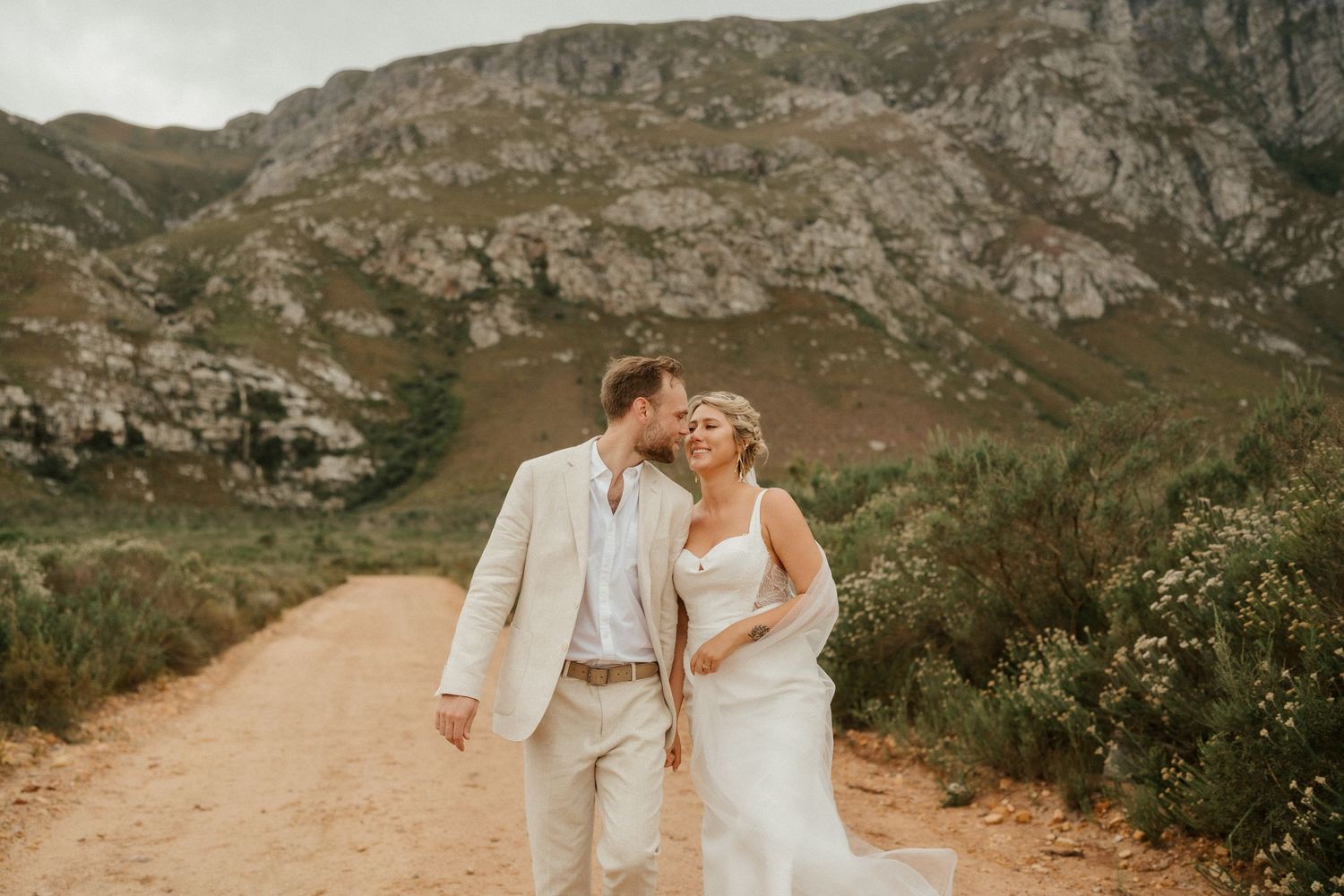 Romantic wedding couple embracing on a dirt path with dramatic mountain landscape behind them.