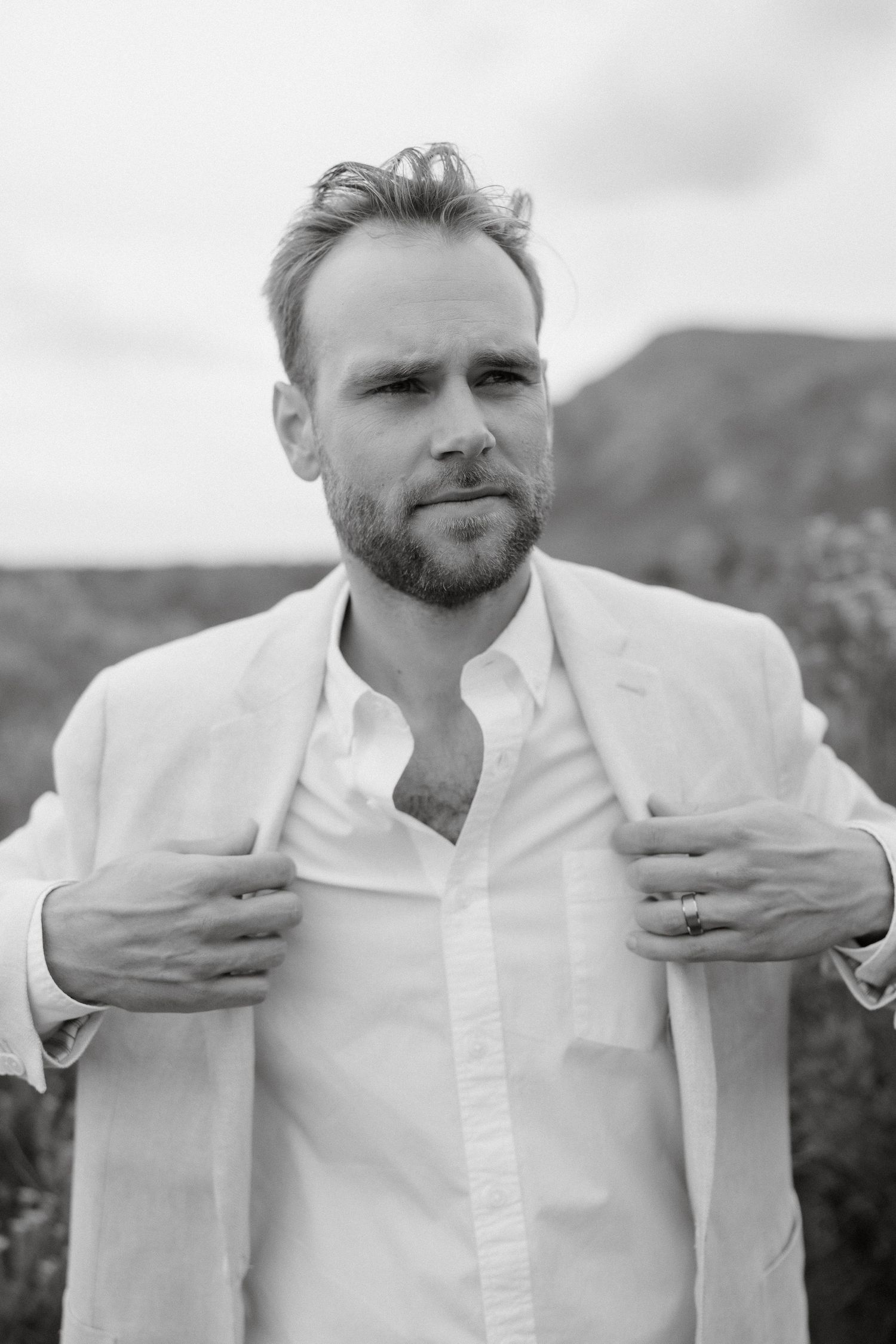 A black and white portrait of someone wearing a white suit against a mountain backdrop.
