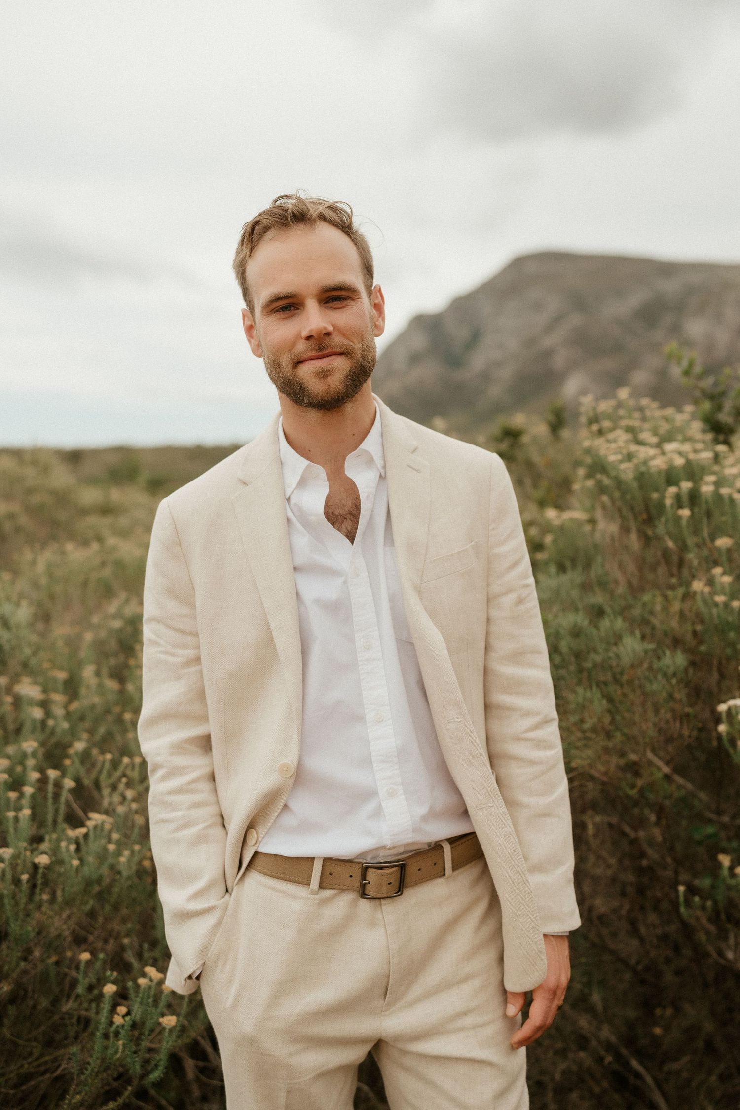 A groom in a cream colored suit and white shirt poses outdoors against a mountain landscape background.