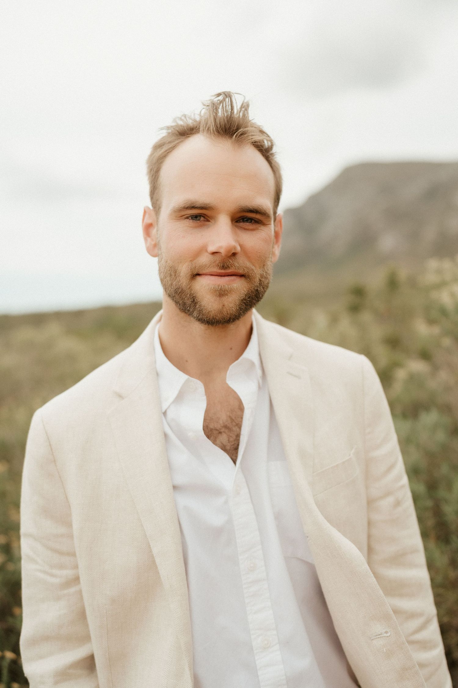 A groom in a cream-colored suit and white shirt poses against mountain scenery.