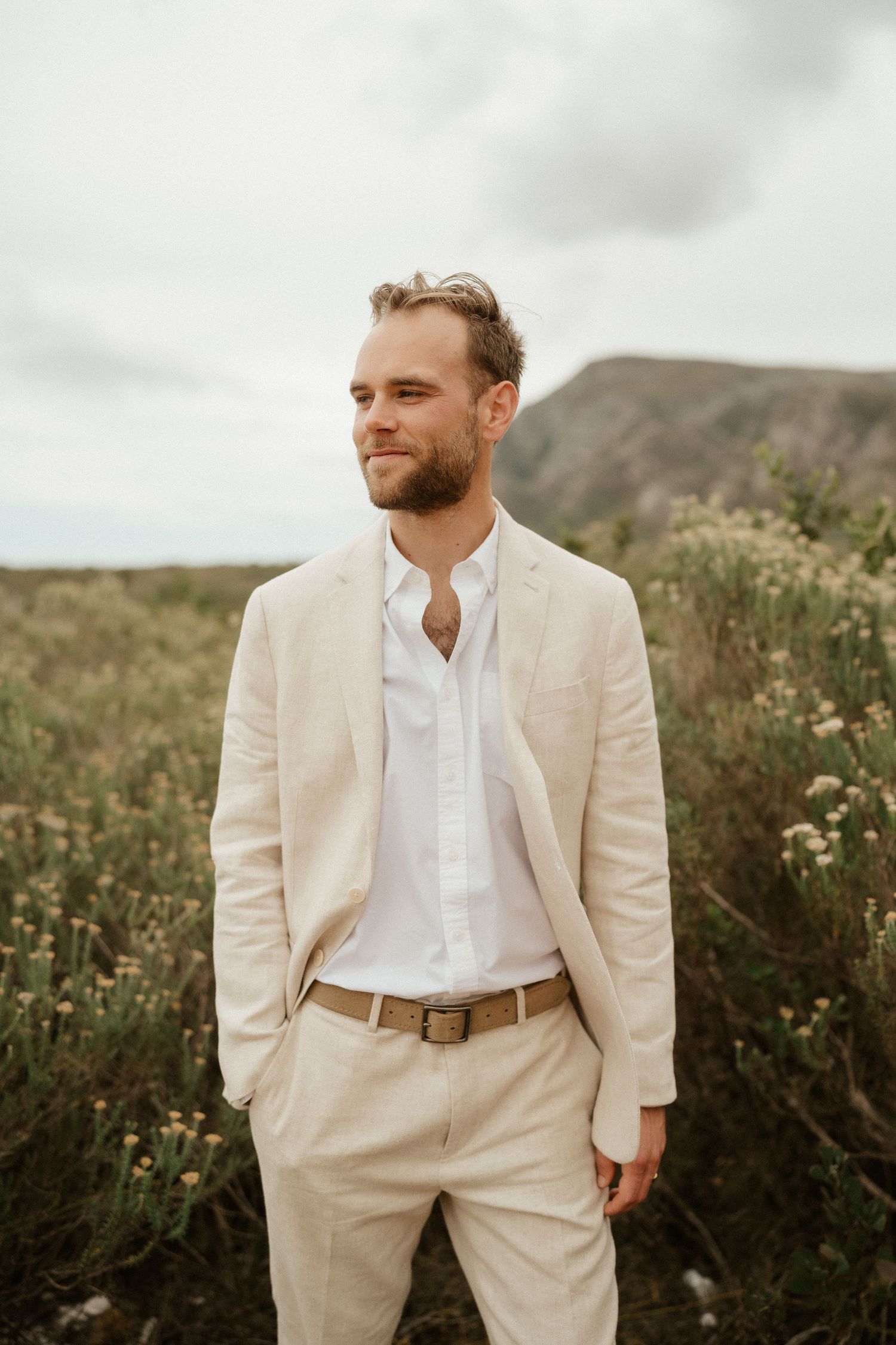 Groom in cream-colored suit stands against mountain landscape backdrop.