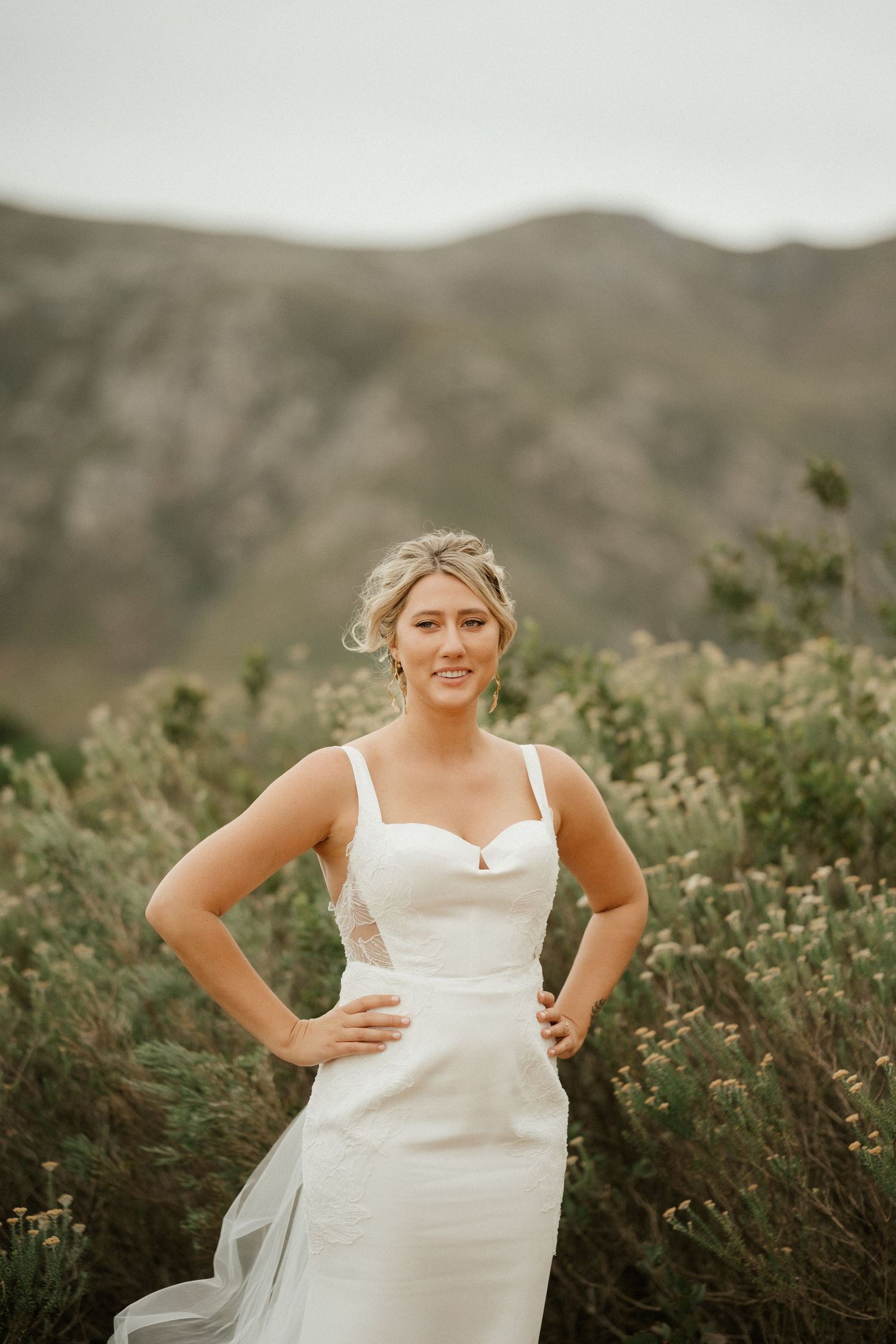 A woman in a white wedding dress poses in a natural outdoor setting with mountains in the background.