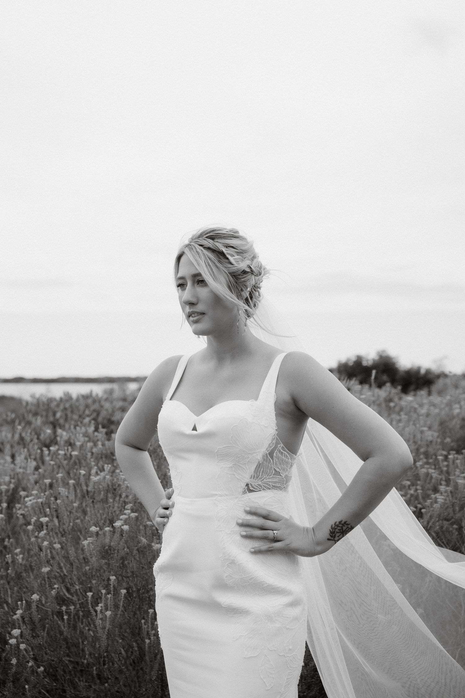 A black and white portrait of a bride in a fitted dress with flowing veil against a natural landscape.