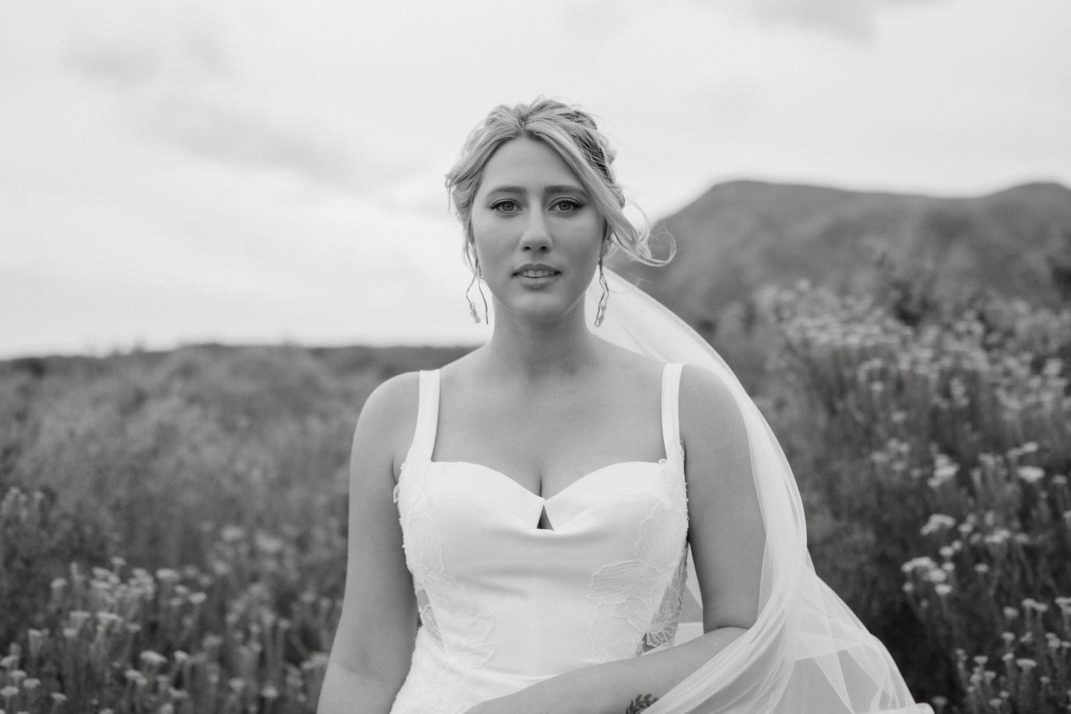 A black and white portrait of a bride wearing a white dress outdoors with mountains and wildflowers in the background.