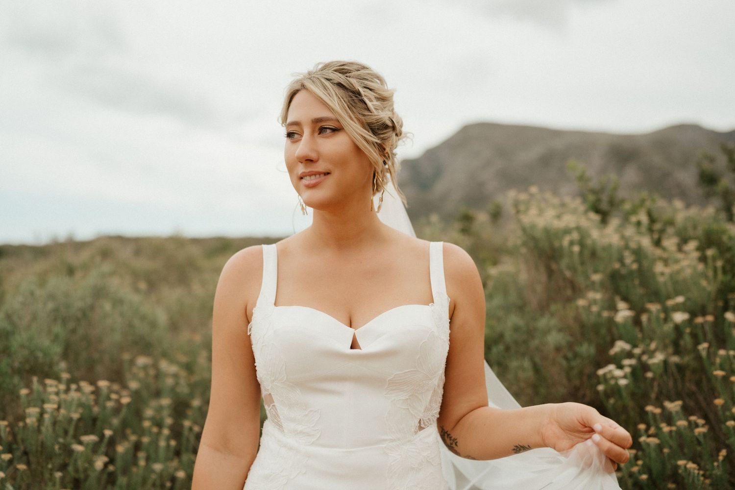 A bride in a white dress poses elegantly in a field with mountains in the background.