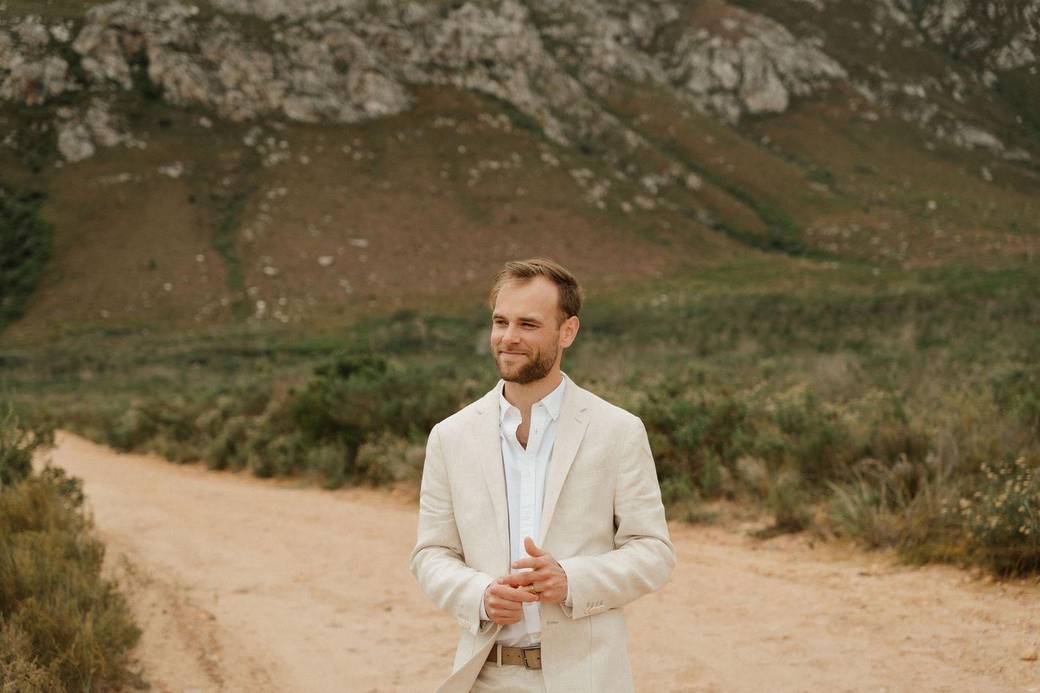 Groom in light colored suit stands thoughtfully on mountain path.