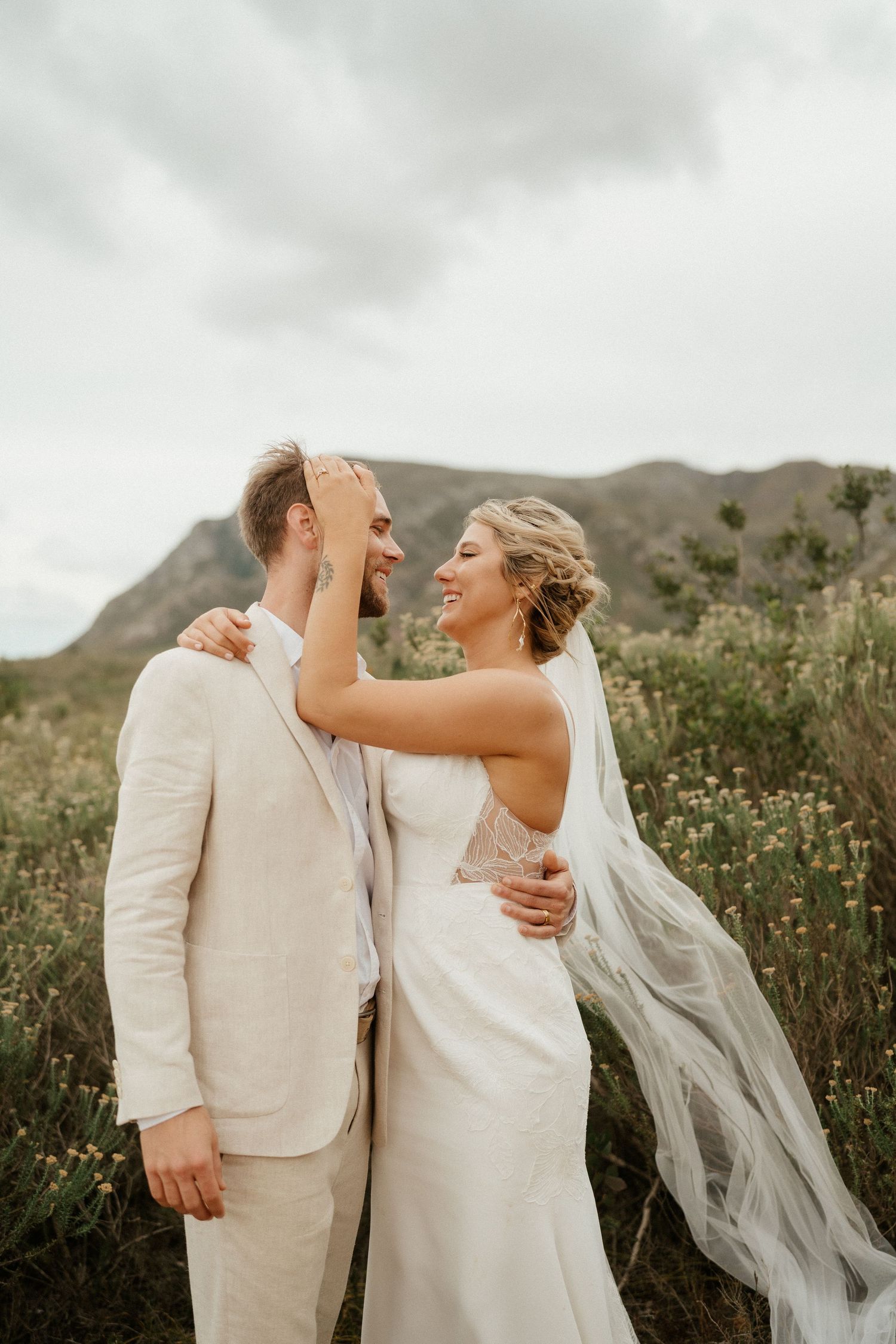 A joyful wedding couple laughs together on a hill with flowing veil and light colored attire against a cloudy sky.