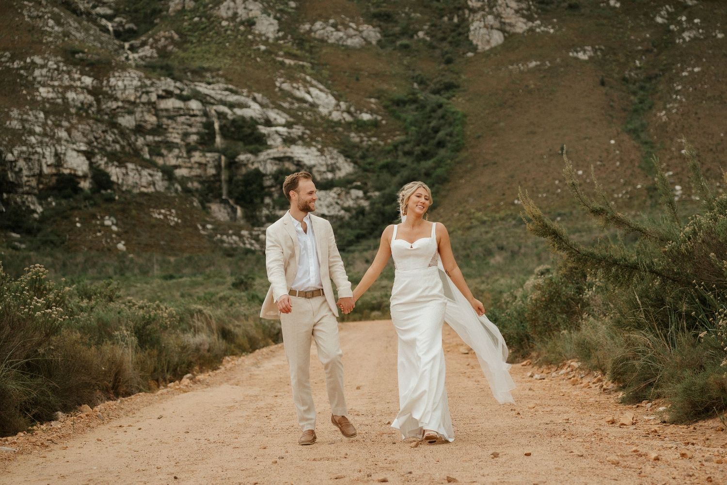 A couple in white wedding attire walking hand in hand on a mountain trail.