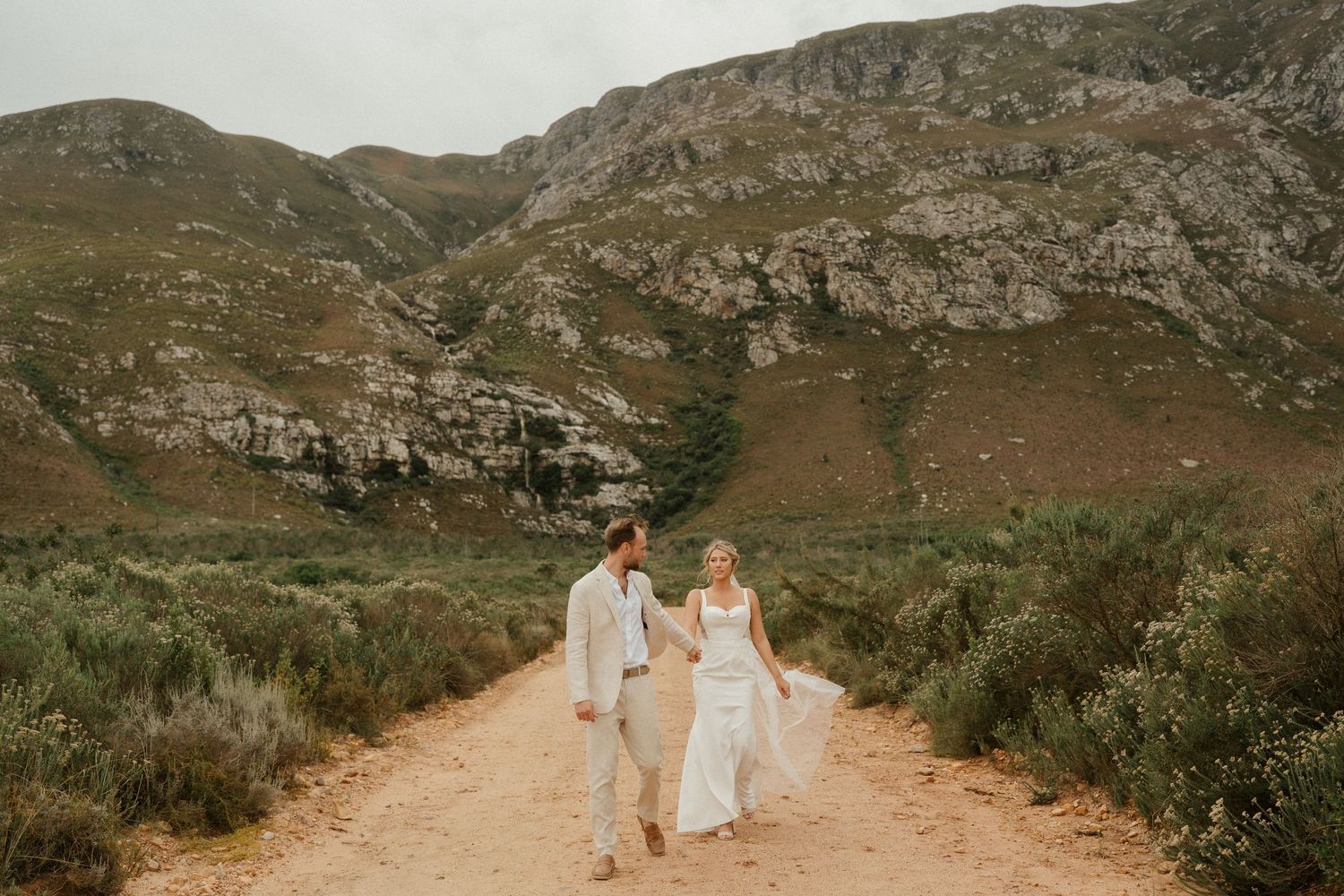 A couple walks hand in hand on a dirt path through rugged mountain terrain during a golden afternoon.