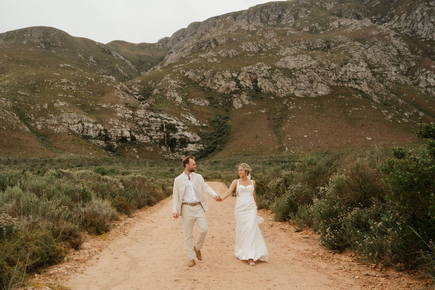 A couple dressed in white stroll down a scenic mountain path in nature.