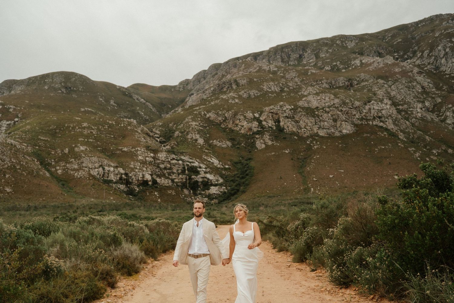 Two people walk hand in hand on a dirt path with dramatic mountain scenery in the background.