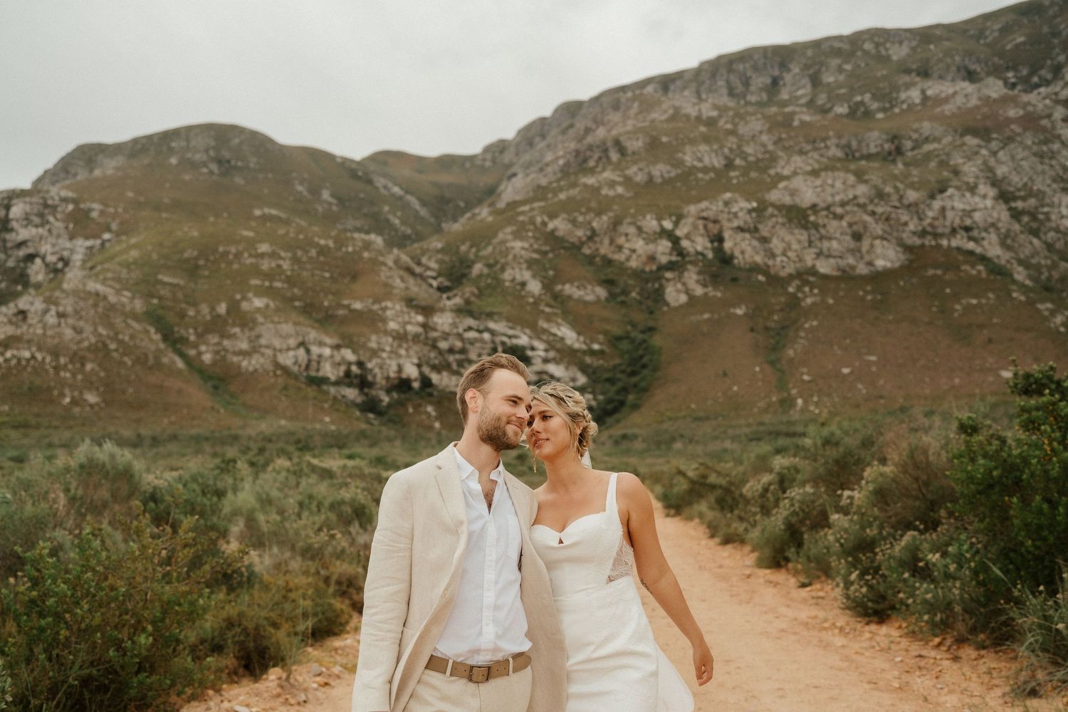 A couple embraces on a dirt path with rugged mountain landscape in the background.