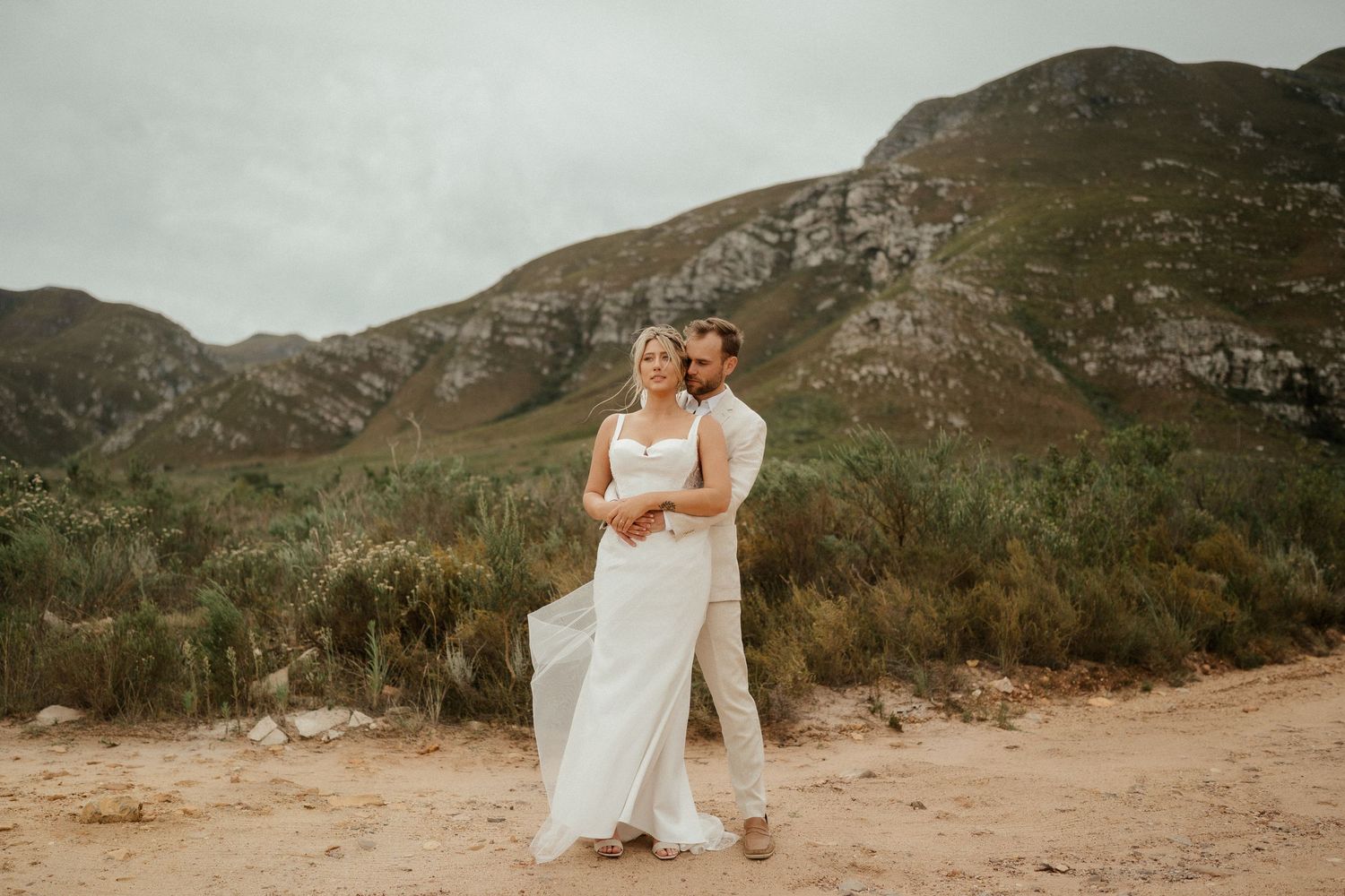 A couple in wedding attire share an intimate moment on a dirt path with mountains in the background.