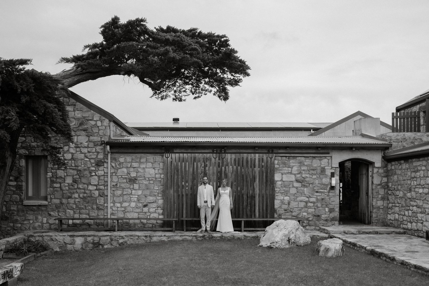 Black and white photo of a stone building exterior with a large tree and two figures standing by the entrance.