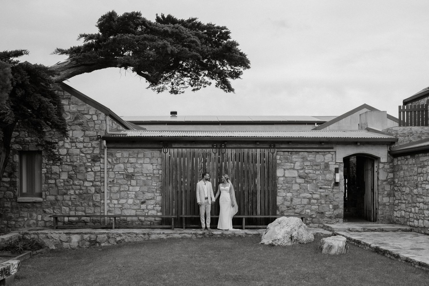 Black and white photo of stone building with wooden gates and cypress tree.