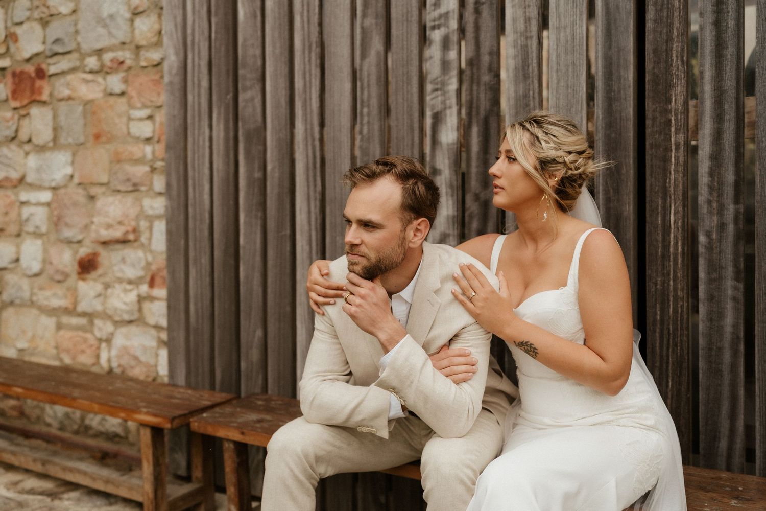 Candid moment of couple sitting on wooden bench by rustic wall.