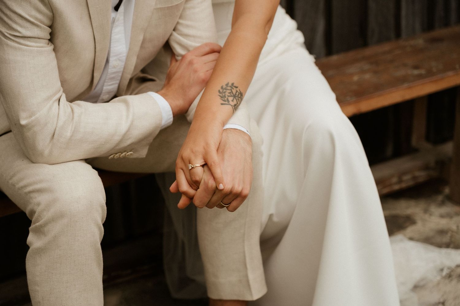 A close-up shot of a couple holding hands while sitting, with the person in a white dress showing a small tattoo.