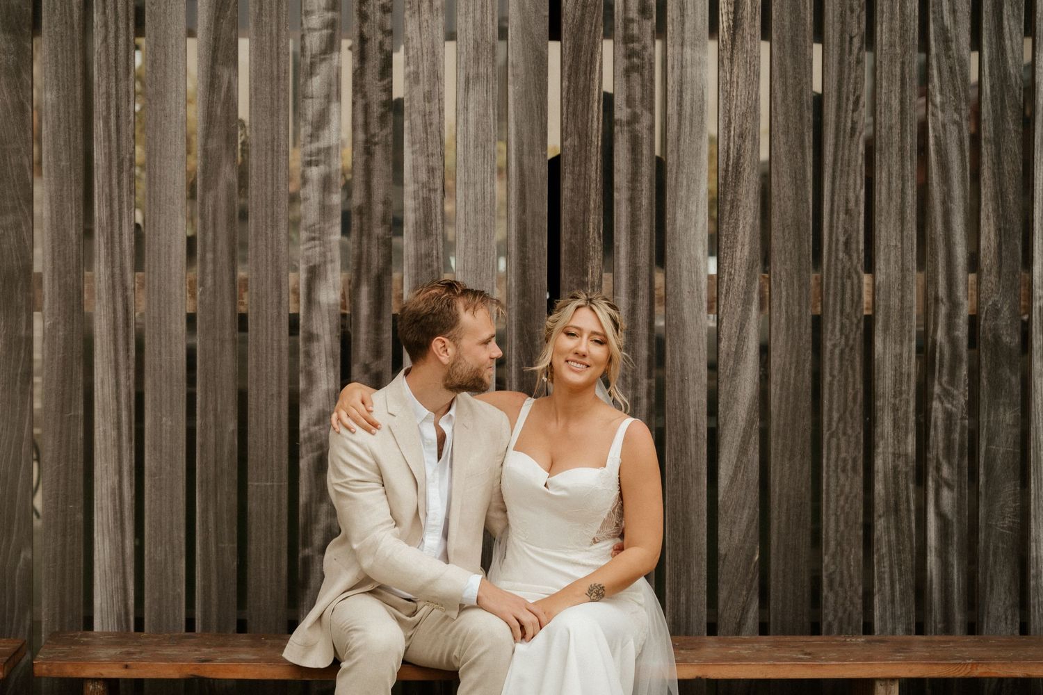A couple sits together on a bench against a rustic wooden fence.