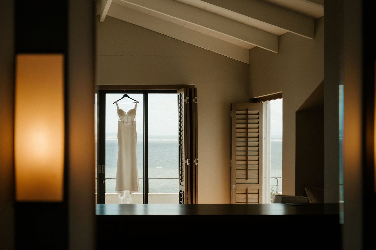 A wedding dress hangs in silhouette against a window with ocean views and warm interior lighting.