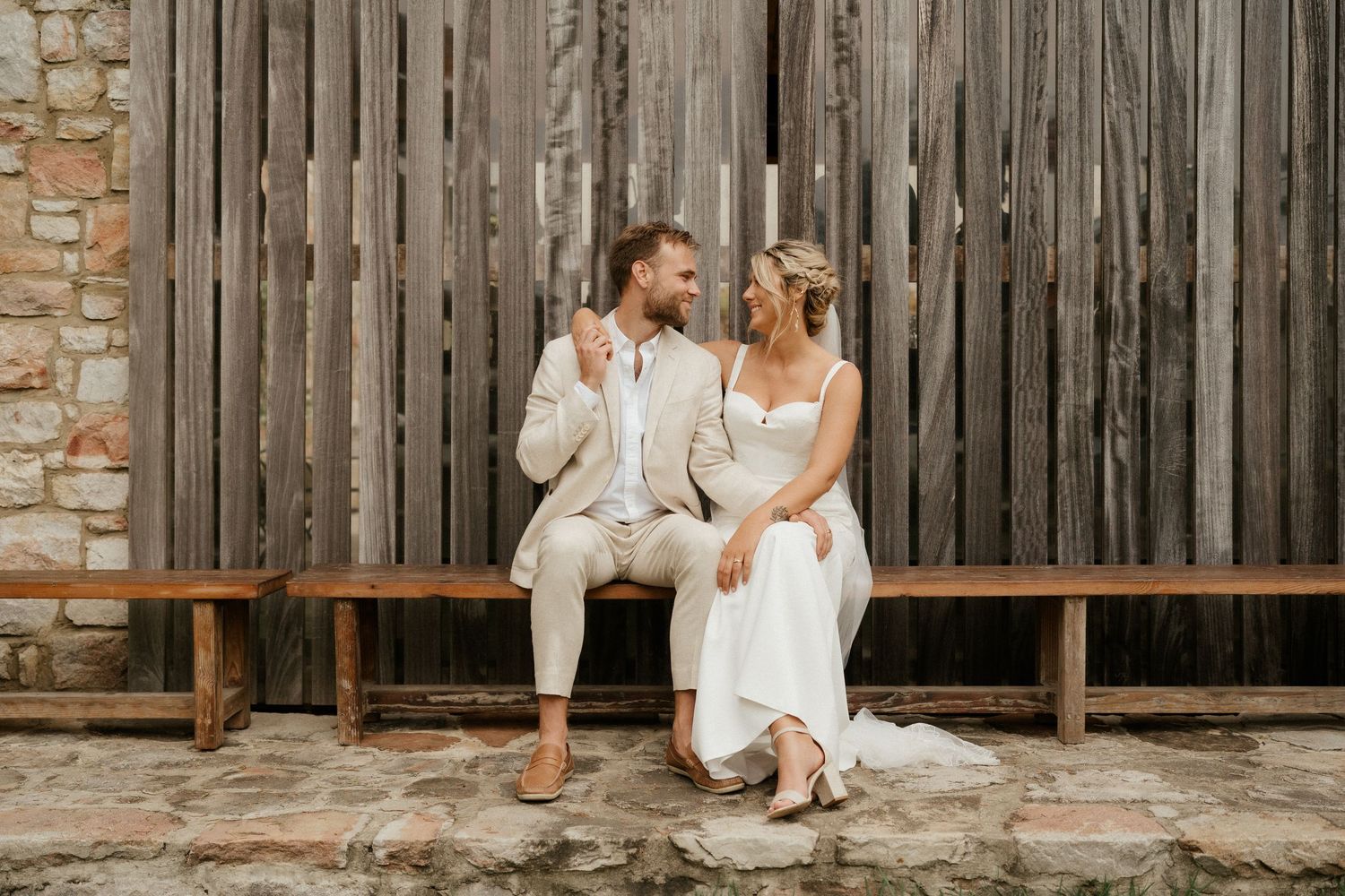A couple in wedding attire sits together on a wooden bench against a rustic barn wall backdrop.