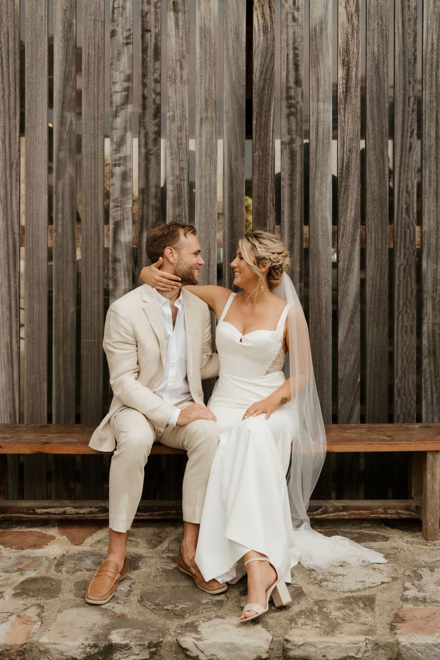 Newlyweds share an intimate moment while sitting on a wooden bench against a rustic slatted wall.