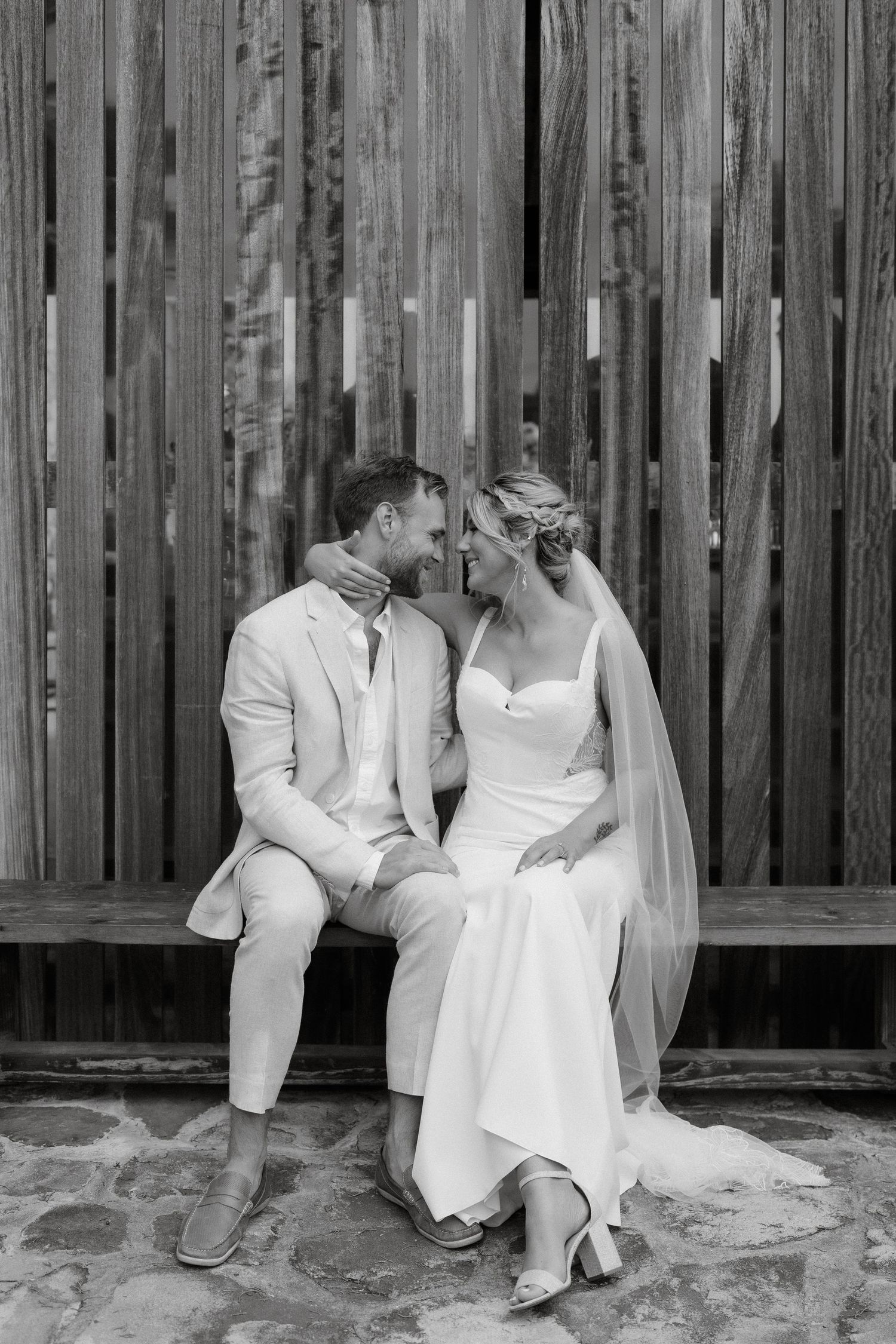 A romantic black and white wedding photo of a couple sitting on a bench against a rustic wooden fence.