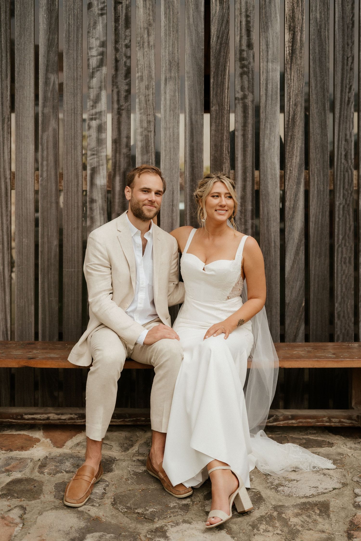A couple in wedding attire sits together on a wooden bench against a rustic wooden wall background.