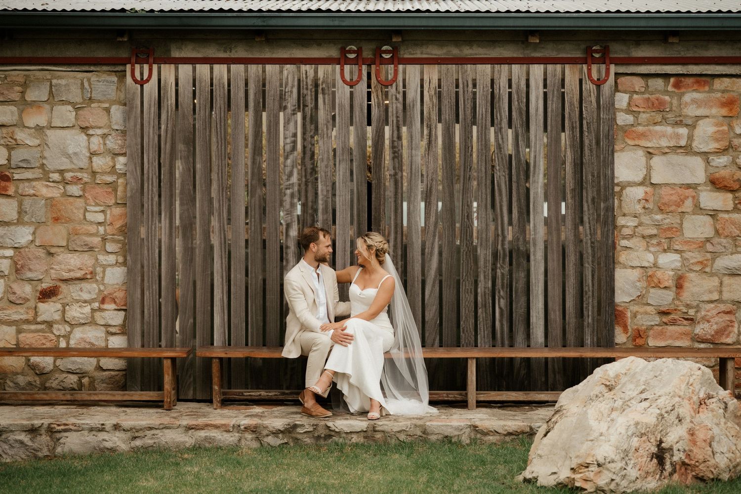 A romantic wedding couple sits on a bench by a rustic wooden gate and stone wall.