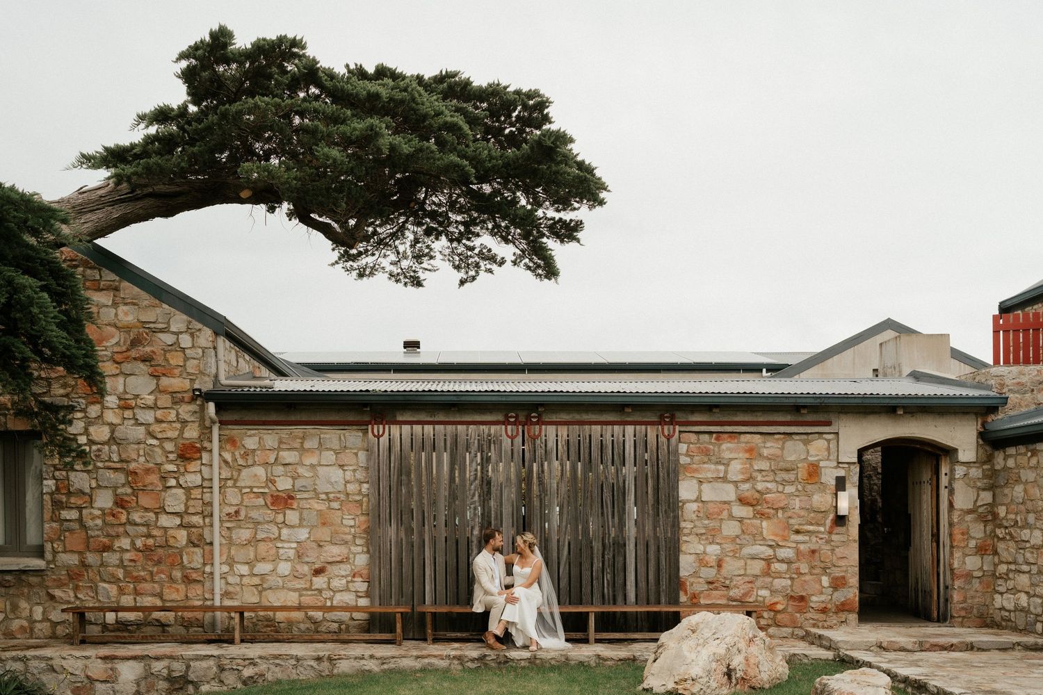 A rustic stone building with wooden doors and an overhanging tree creates a charming wedding venue setting.