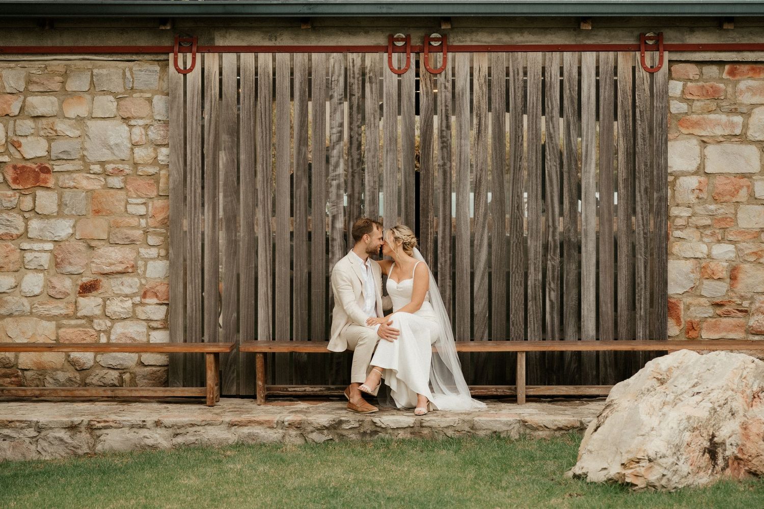 A newlywed couple shares an intimate moment on a wooden bench against a stone and wood barn wall.