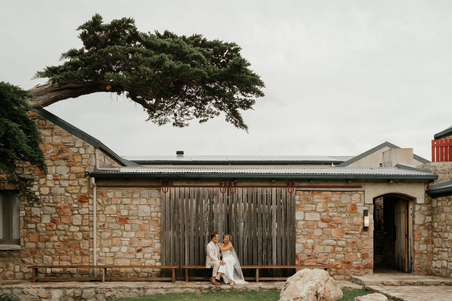 Person sits on rustic stone wall bench beside weathered wooden door and tree at historic building.