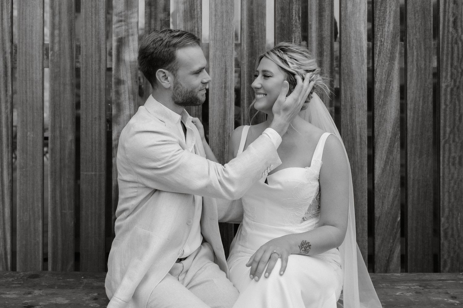 A tender black and white moment between two people in white attire sharing an intimate gesture against a wooden fence.