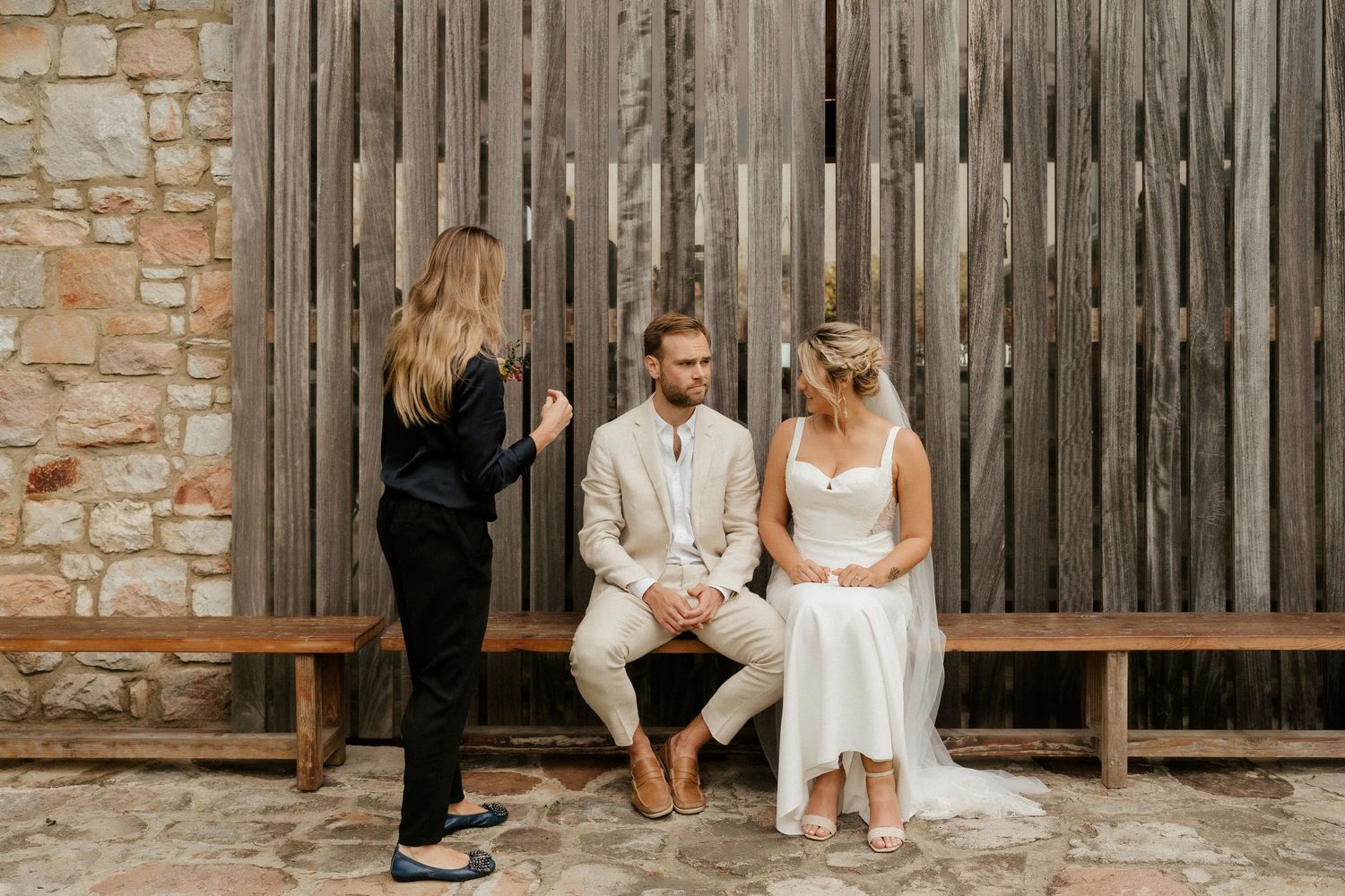 A couple sits together on a rustic wooden bench against a weathered fence while talking with someone in black.