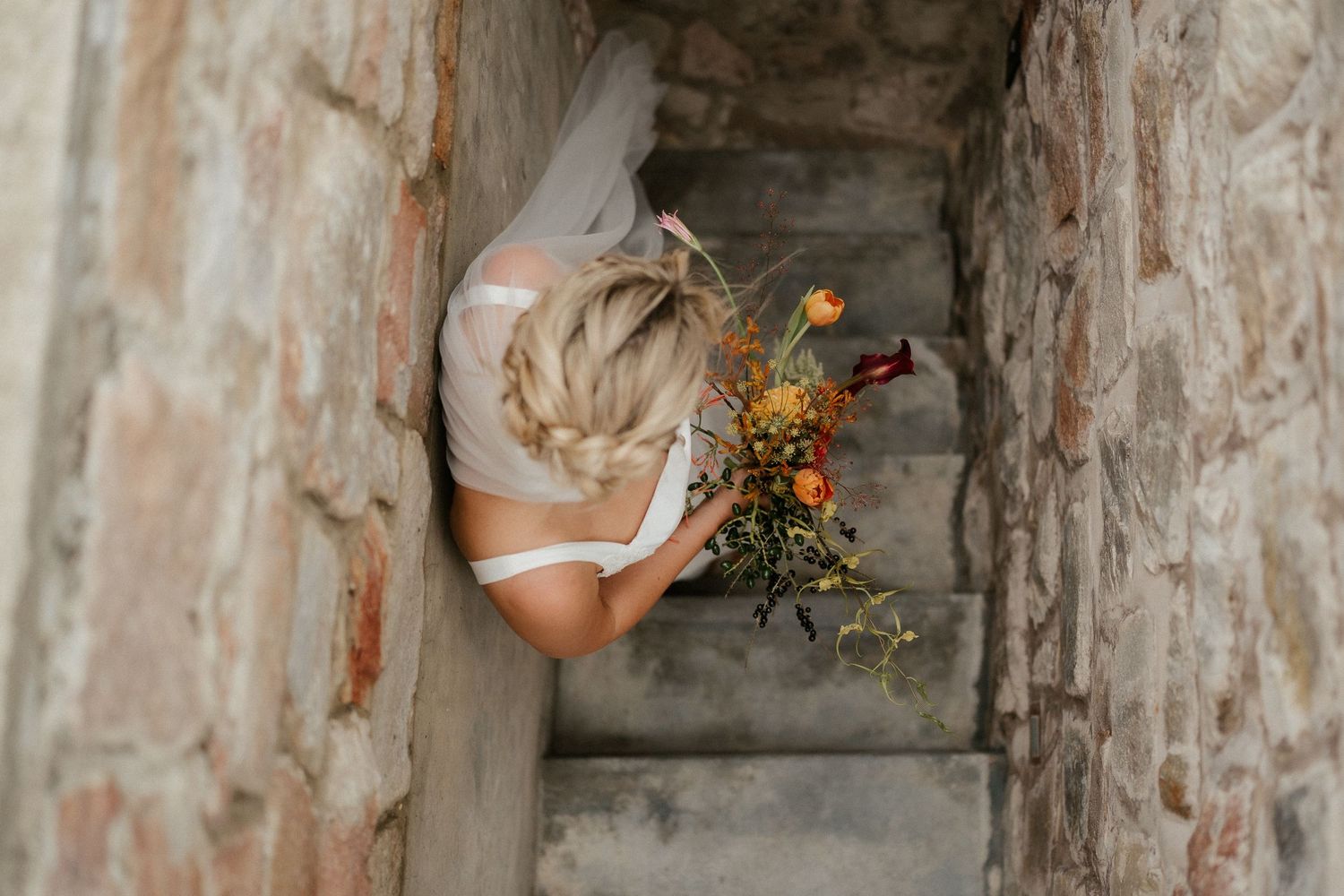 A bride in a white dress holds a bright yellow wildflower bouquet while descending stone stairs.