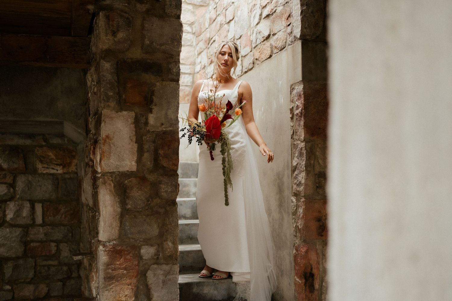 A figure in an elegant white wedding gown poses on rustic stone steps beside an old stone wall.