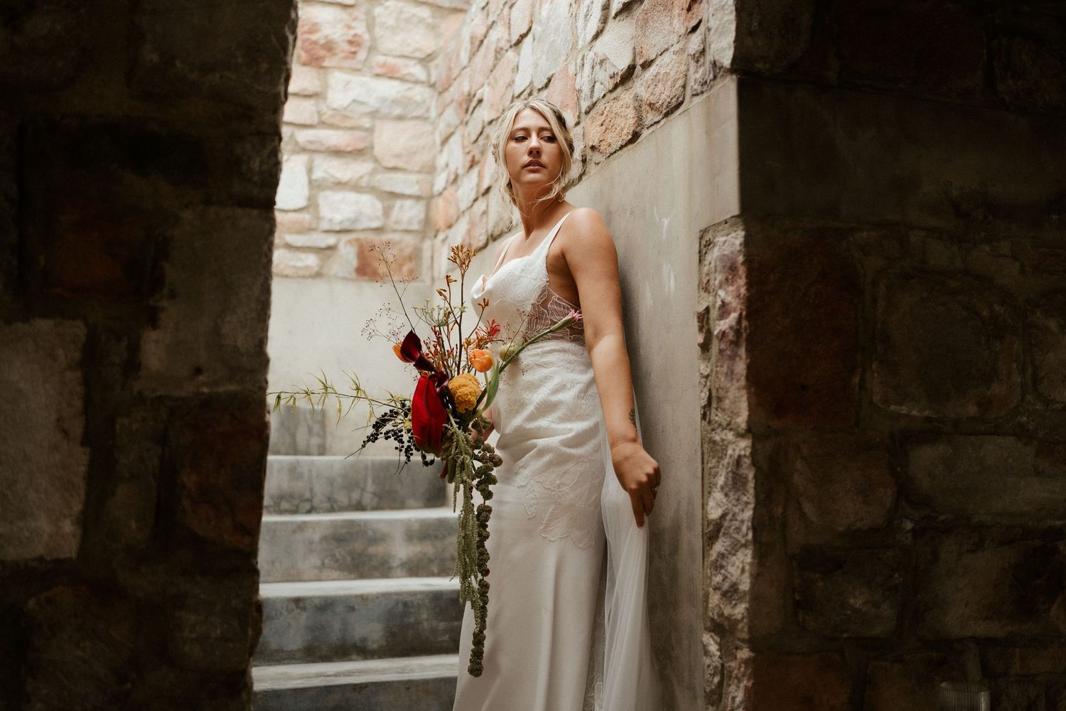 An elegant wedding dress silhouette against stone walls with a colorful bouquet.