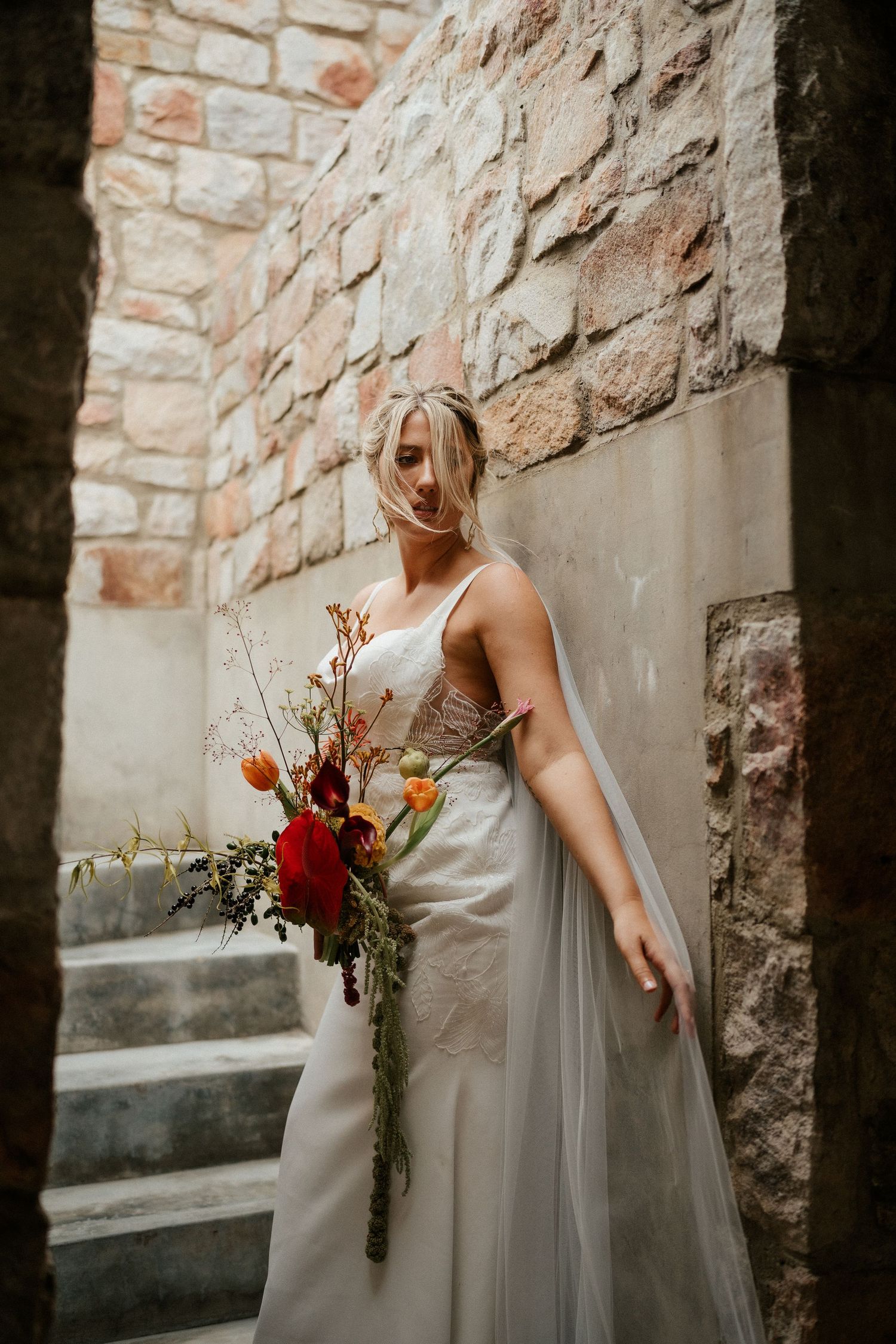 Wedding scene with a white gown against stone walls holding an autumn-colored bouquet.